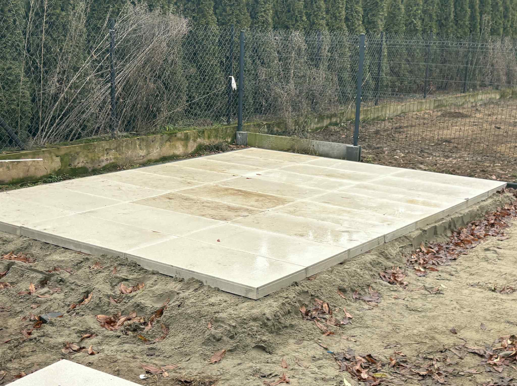 Ułożona, jasna kostka brukowa na piasku, widok z góry. W tle siatka ogrodzeniowa i zielone drzewa. Świeżo wykonana podbudowa pod taras lub ścieżkę.