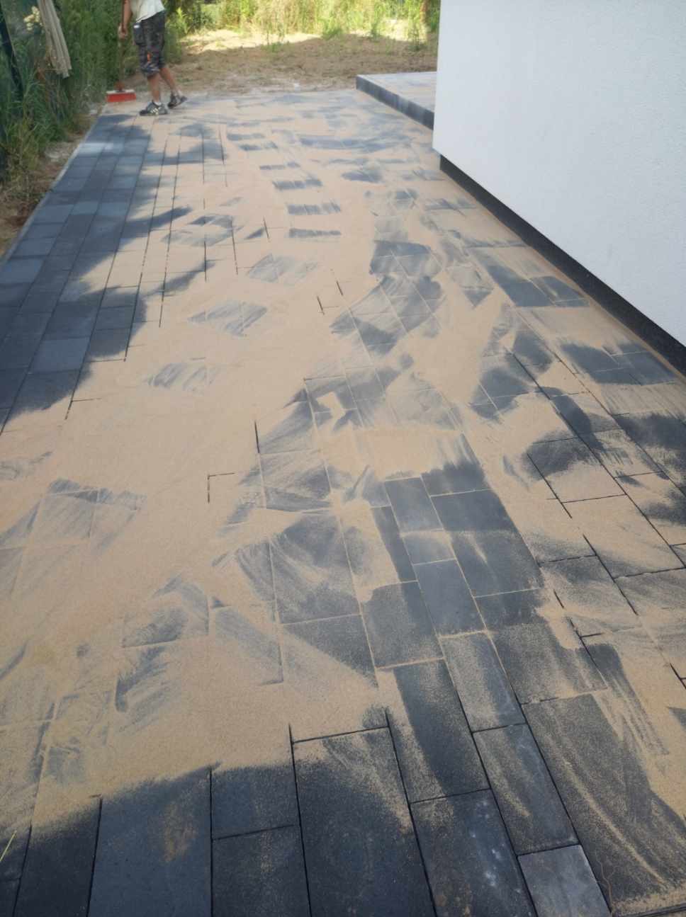 Ciemnoszara kostka brukowa zasypana piaskiem kwarcowym, przygotowana do zagęszczenia. Widoczny fragment budynku z białą elewacją i osoba z miotłą w tle.