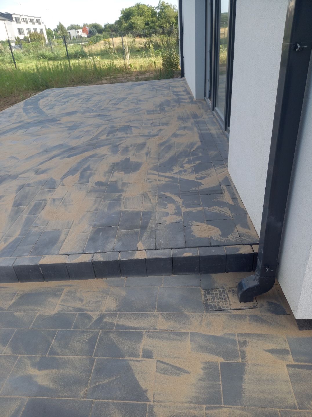Nowoczesny taras z szarej kostki brukowej, zasypanej piaskiem, z czarną rynną przy białej ścianie budynku. Widok z bliska na fragment tarasu.