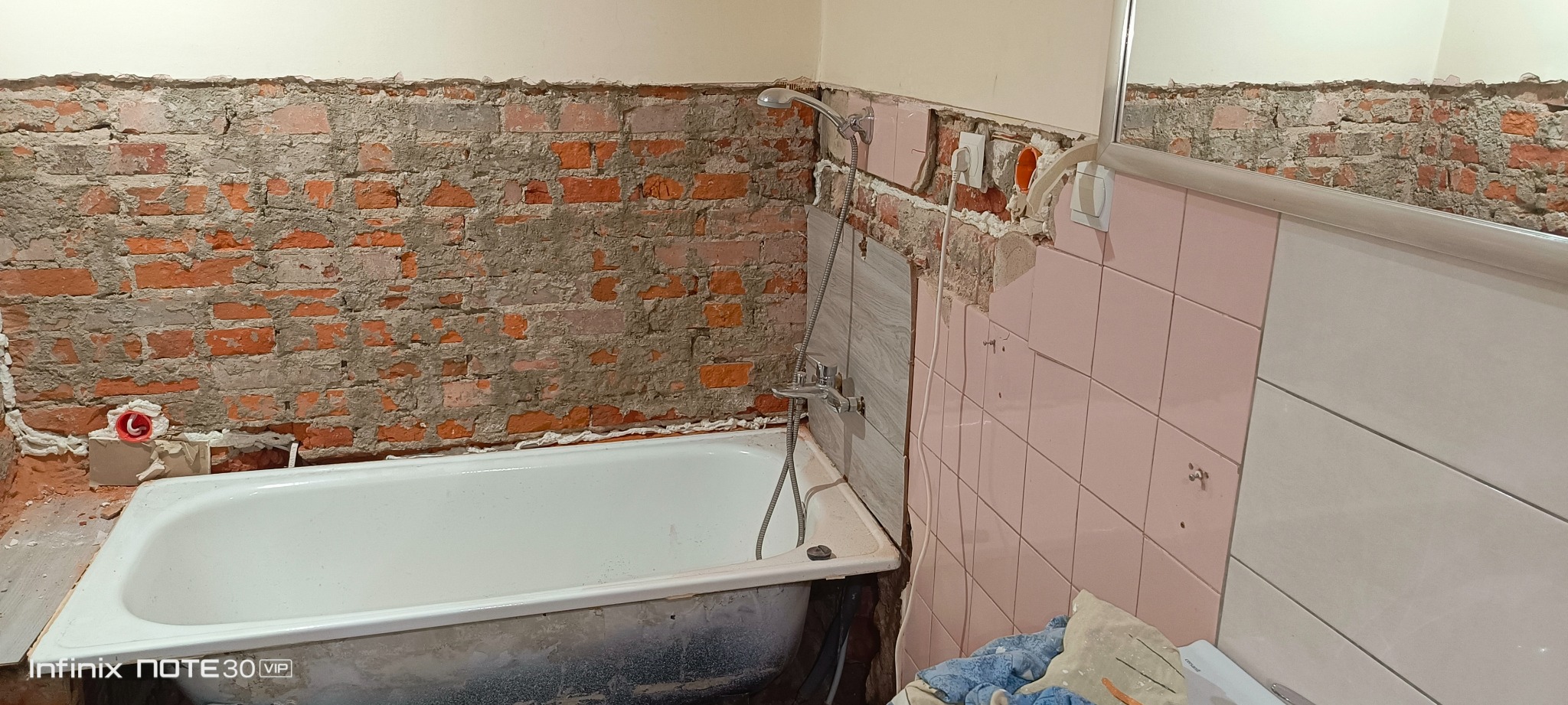 Łazienka w trakcie remontu: odsłonięta cegła, częściowo usunięte różowe płytki, widoczna wanna i instalacja wodna. Etap prac przed wykończeniem.