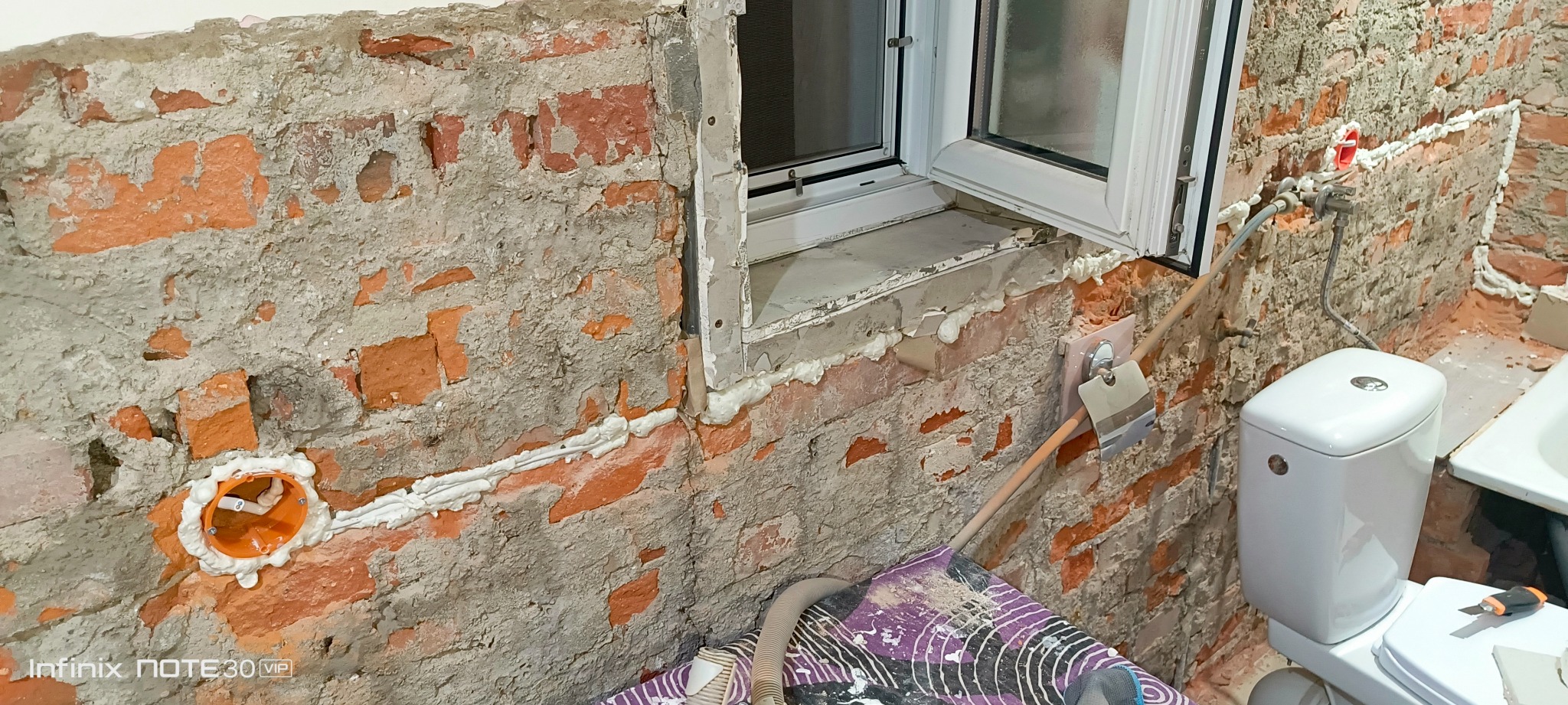Ceglana ściana w trakcie remontu łazienki z nowym oknem, instalacją elektryczną i sanitarną. Widoczny otwór na gniazdko, rury i częściowo zamontowana toaleta.