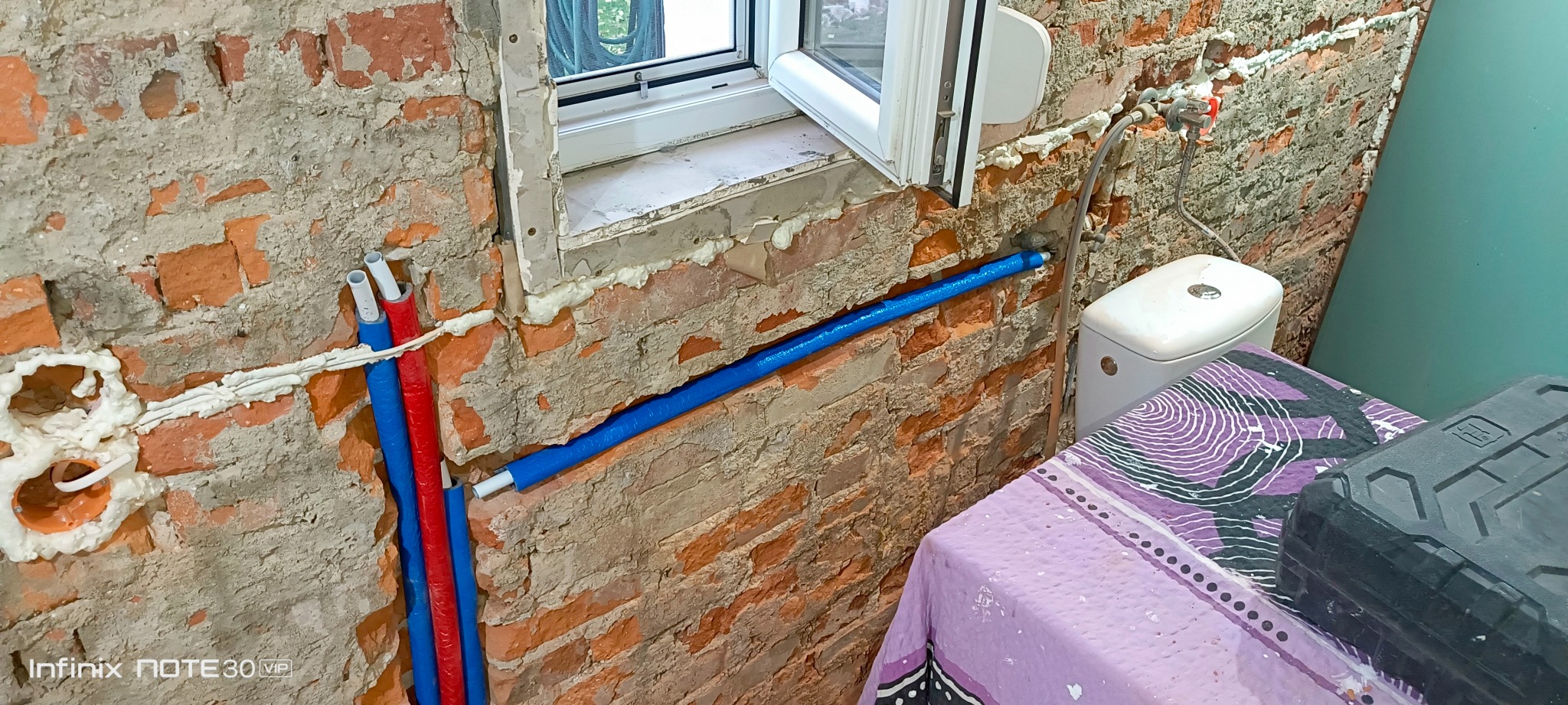 Czerwono-niebieskie rury instalacji wodnej w surowej, ceglanej ścianie łazienki, z widocznym oknem i fragmentem toalety. Pianka montażowa wokół rur.