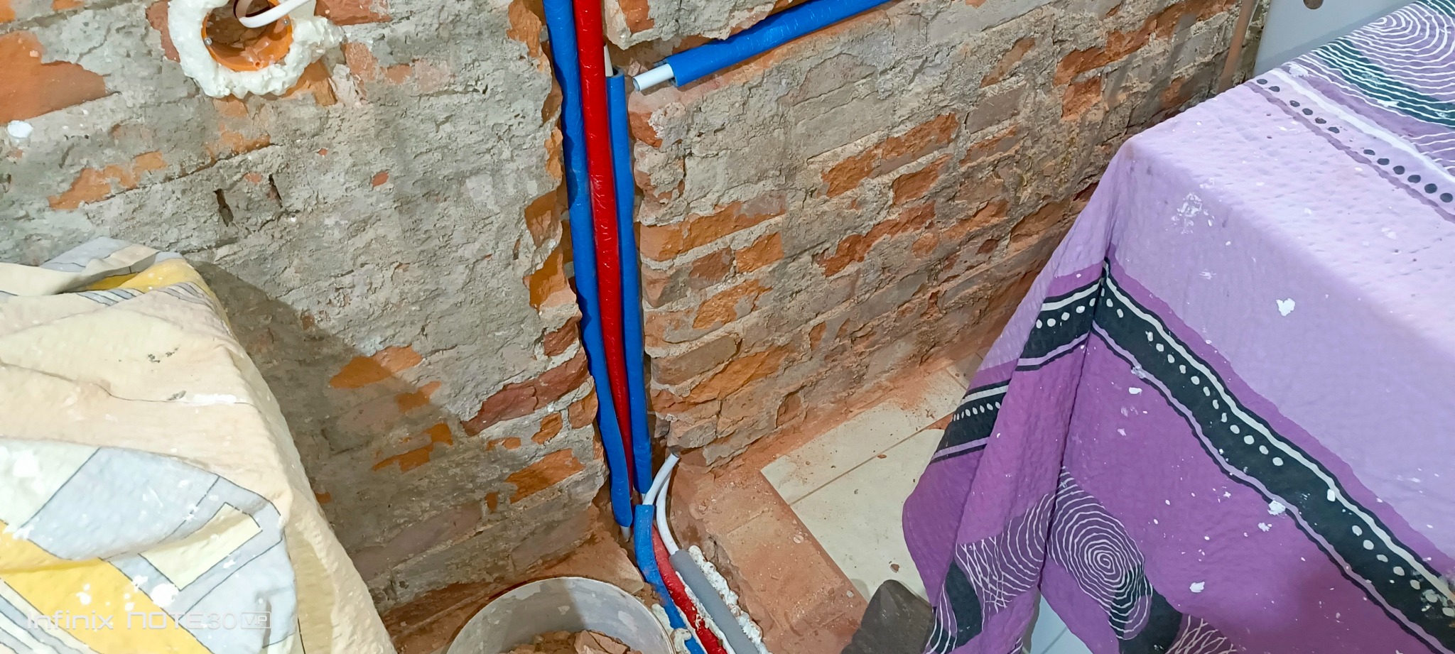Instalacja rur w bruzdach ceglanej ściany, zabezpieczonych pianką montażową. Widoczny fragment podłogi i przykryty mebel. Prace hydrauliczne w trakcie remontu.