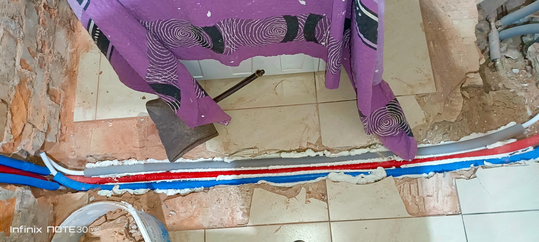 Instalacja rur w bruzdach w podłodze, zabezpieczonych pianką montażową. Widoczne rury w kolorach: niebieskim, czerwonym i szarym. Fragment podłogi z płytek i ściany z cegły.