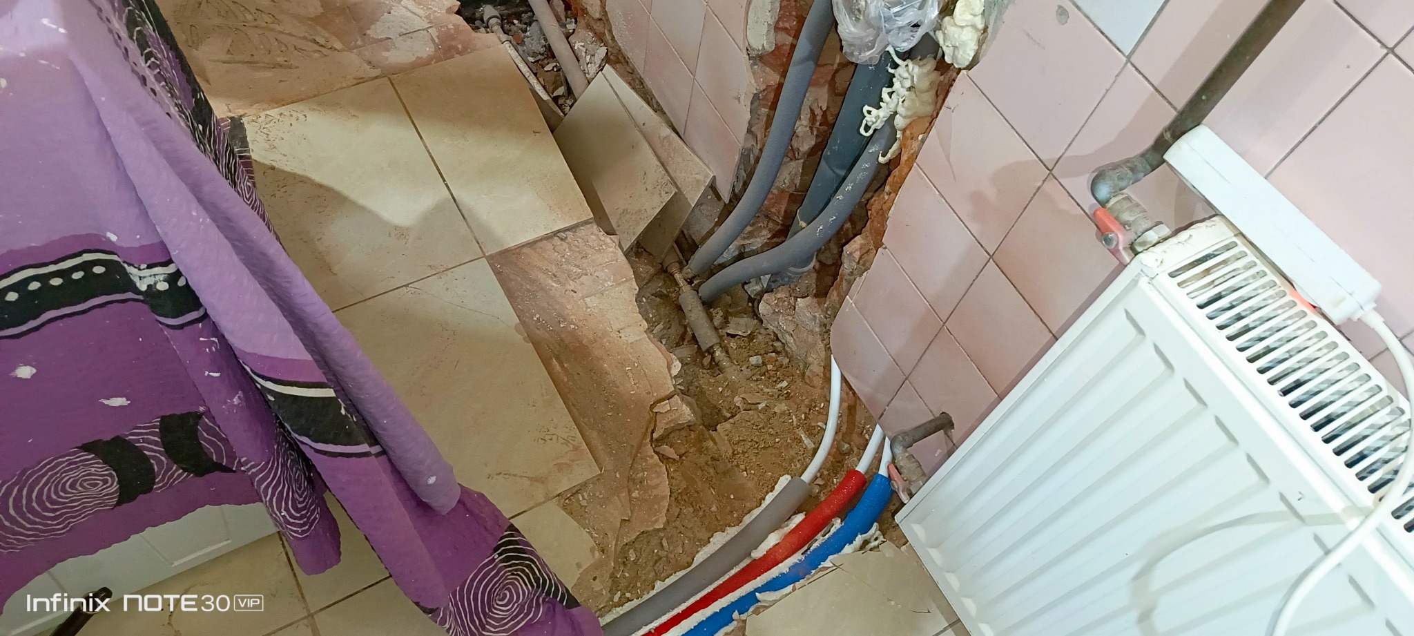 Odsłonięte rury w ścianie łazienki podczas remontu, widoczne fragmenty instalacji grzewczej i hydraulicznej, grzejnik, fragment zasłony w lewym rogu.