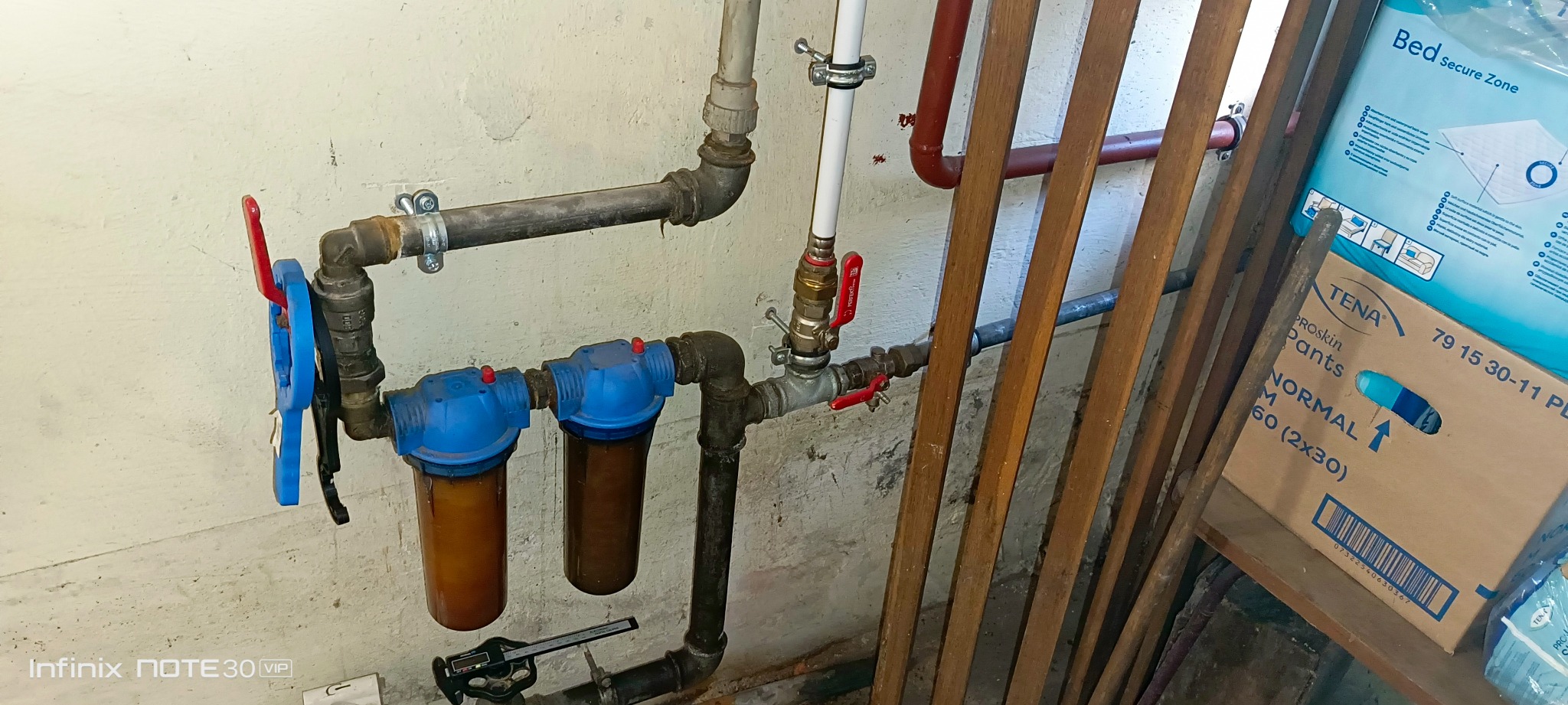 Kompleksowa instalacja wodna z filtrami i zaworami, widoczne rury, złączki i filtry z brązowym osadem. Elementy instalacji przymocowane do ściany.