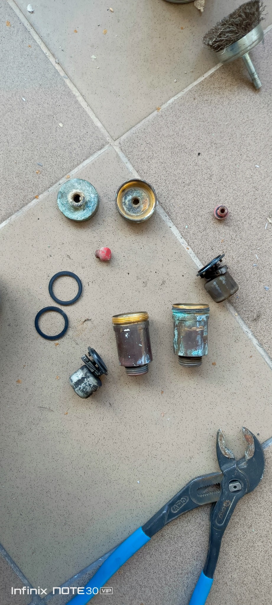 Rozłożone elementy armatury, uszczelki, szczotka druciana i kombinerki na płytkach podłogowych. Widoczne ślady korozji na elementach.