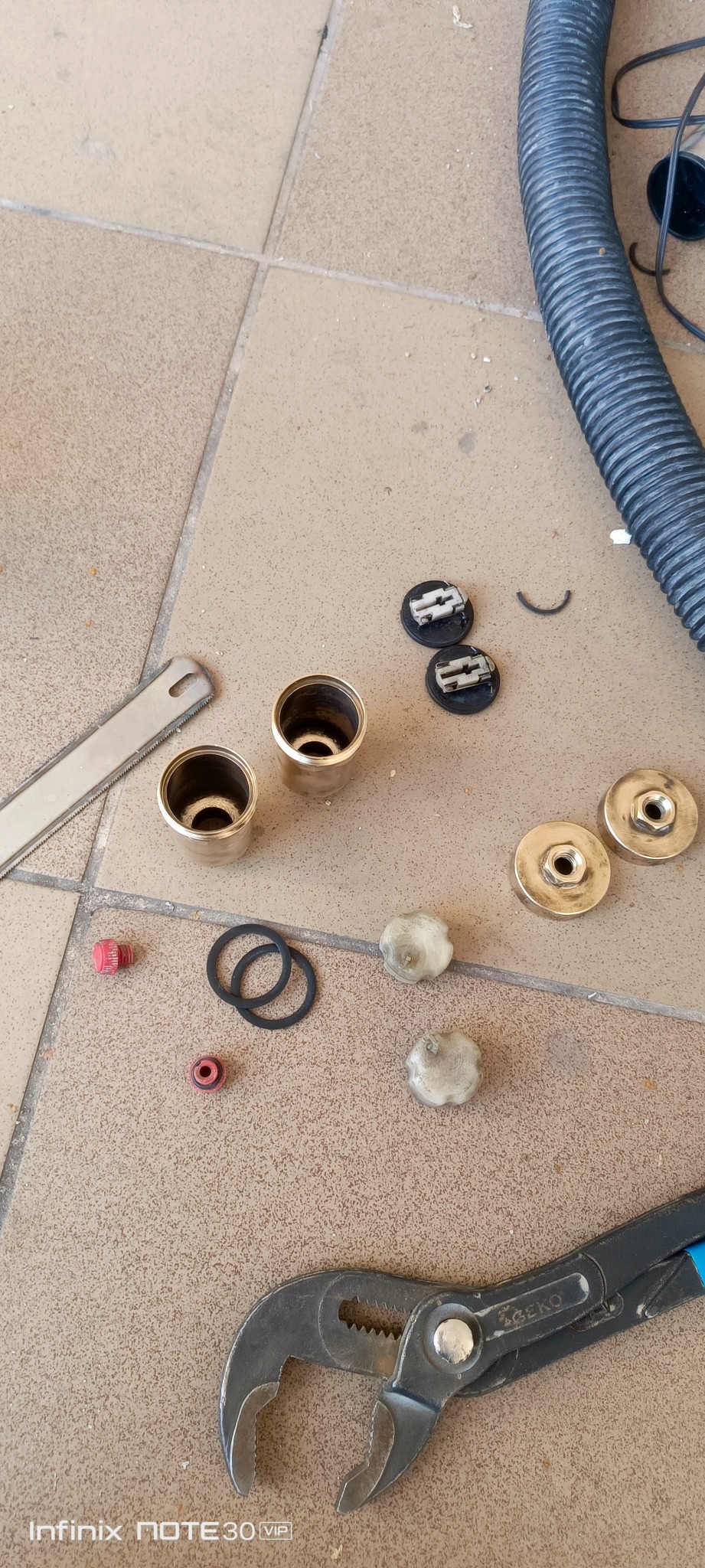 Rozłożone elementy armatury (uszczelki, zawory, nakrętki) i narzędzia (klucz nastawny, piła) na beżowej podłodze z płytek. Przygotowanie do montażu lub naprawy.