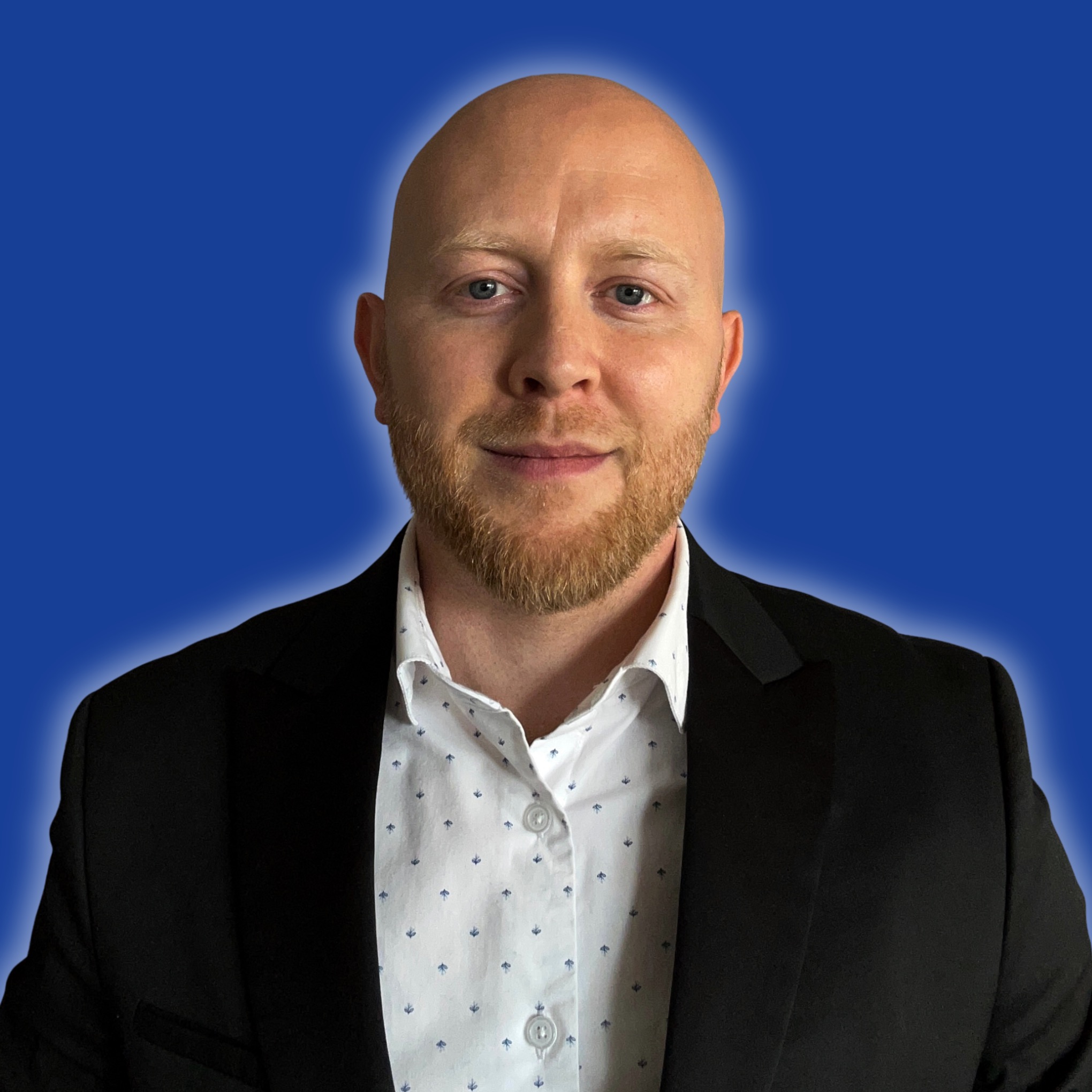 Portret uśmiechniętego, łysiejącego mężczyzny z rudą brodą w czarnej marynarce i białej koszuli na jednolitym, niebieskim tle z delikatną aureolą.