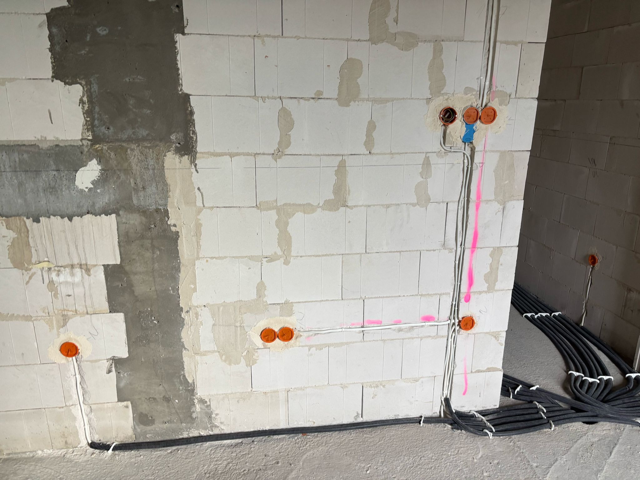 Wewnętrzna instalacja elektryczna w surowym budynku z białych bloczków, widoczne kable w peszlach i pomarańczowe puszki pod gniazdka, ściany częściowo otynkowane.