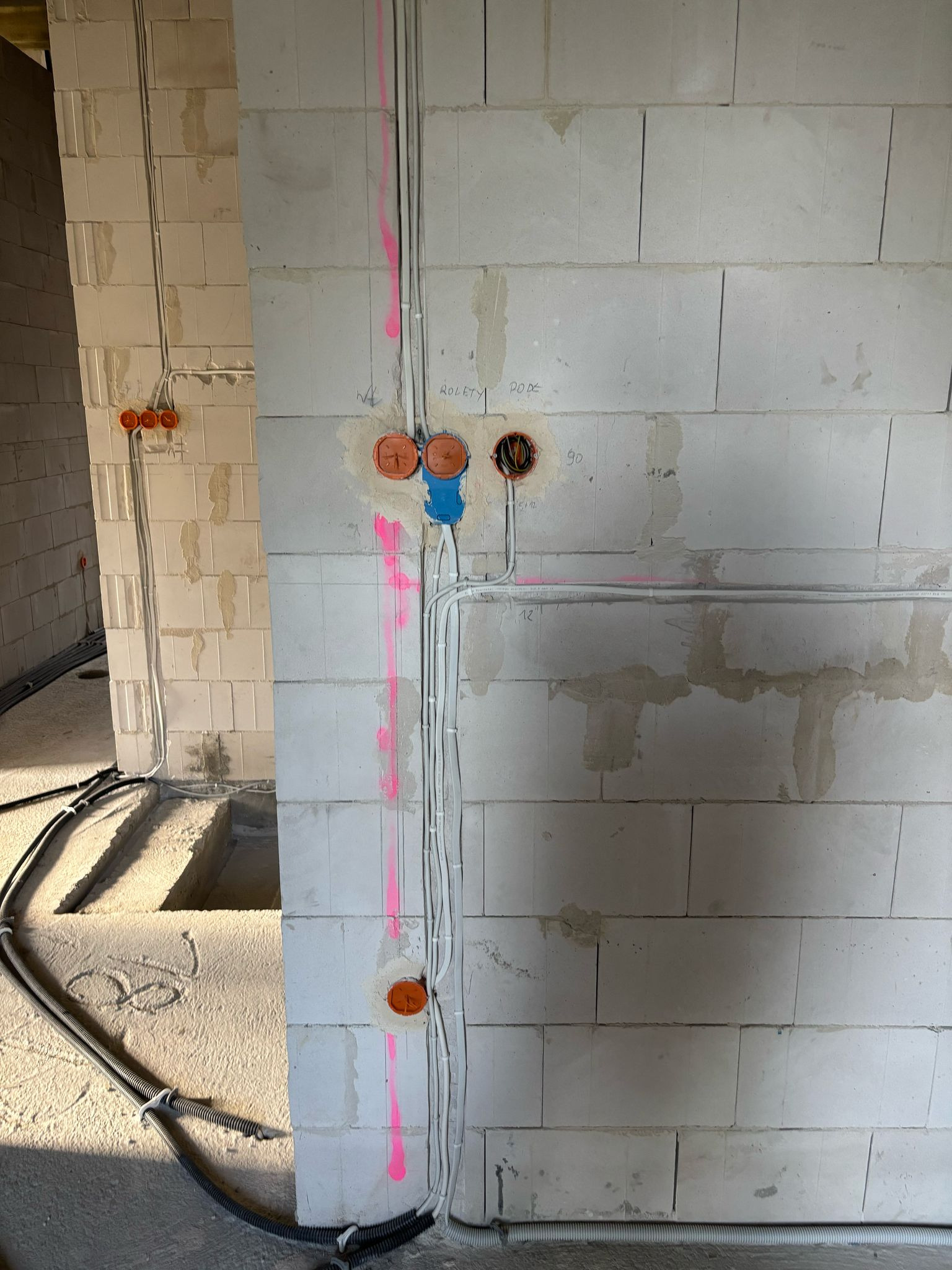 Instalacja elektryczna w surowym budynku z białych bloczków. Widoczne kable w peszlach, puszki instalacyjne, oznaczenia różowym sprayem. Etap prac przed tynkowaniem.