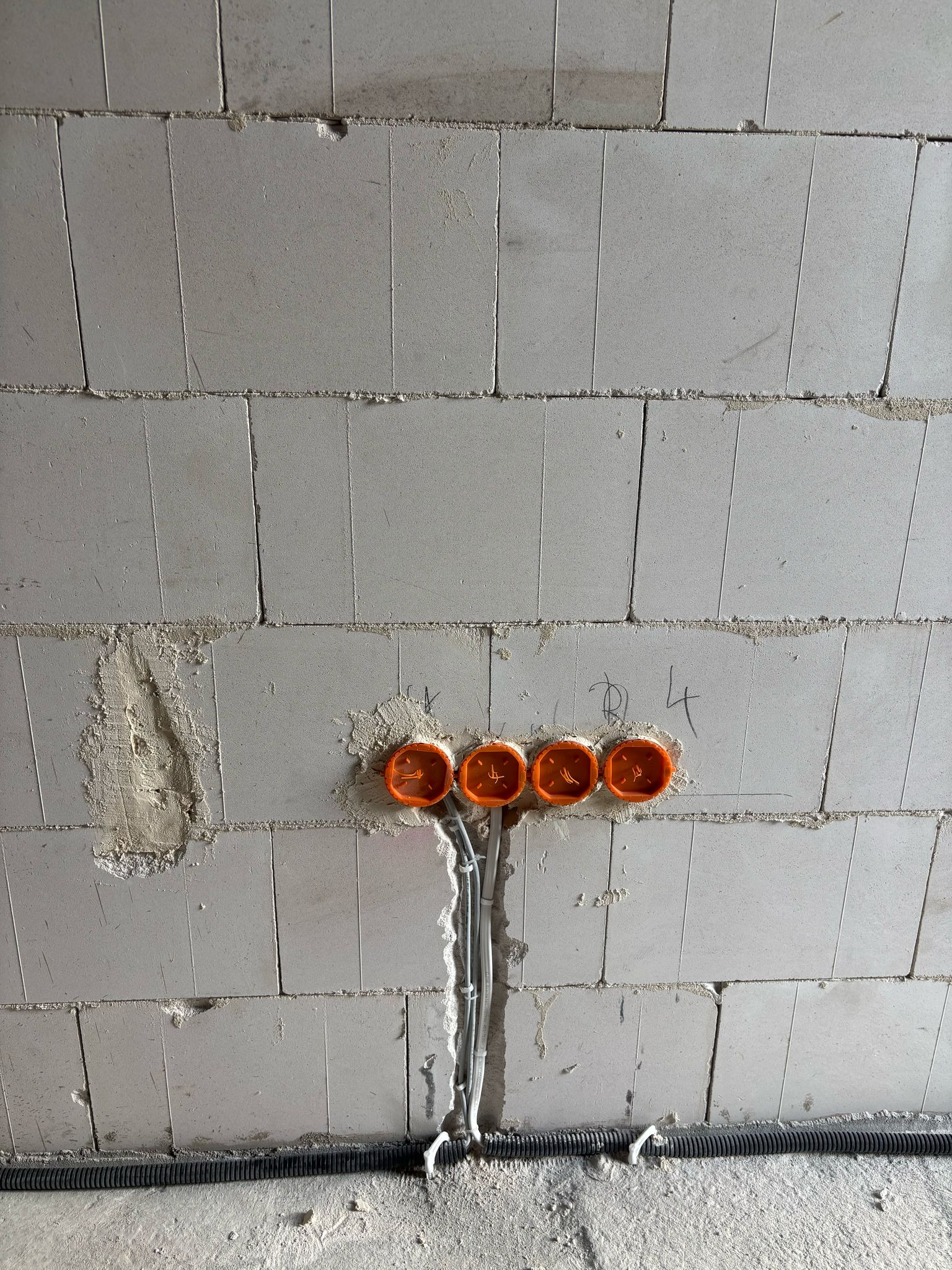 Surowa ściana z betonu komórkowego z zamontowanymi, pomarańczowymi puszkami elektrycznymi i przewodami w białych peszlach. U dołu widoczny czarny peszel.