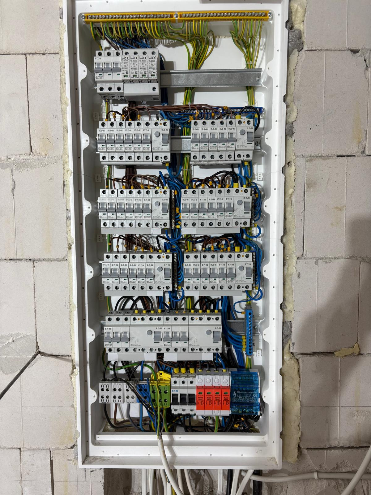 Rozdzielnica elektryczna z zabezpieczeniami w białej obudowie, widoczna na tle ściany z betonu komórkowego w Kleczew. Widoczne przewody i aparatura modułowa.