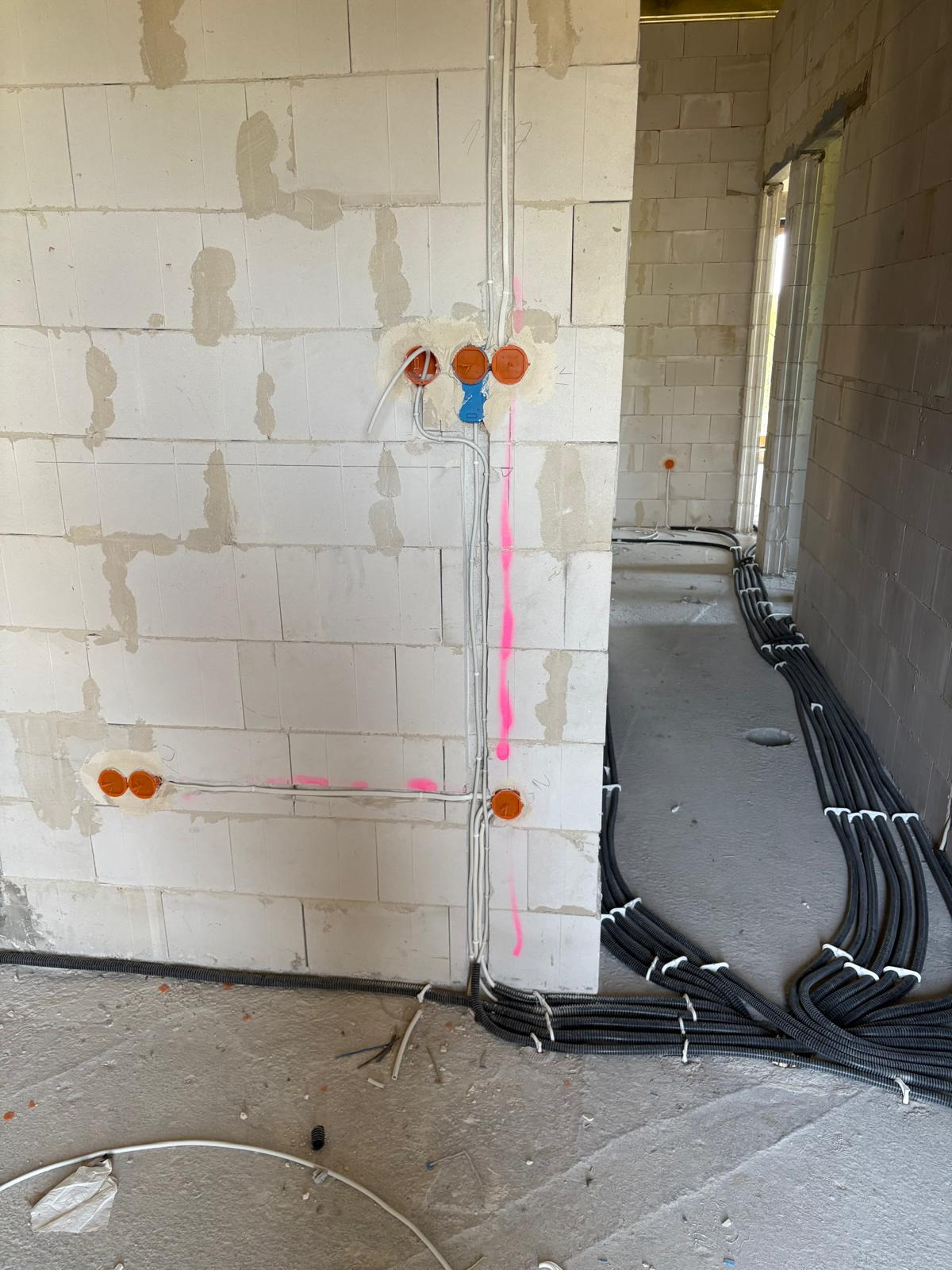 Surowy mur z instalacją elektryczną: pomarańczowe puszki, białe kable, różowe oznaczenia. W tle rury w peszlach na podłodze. Stan surowy budynku.