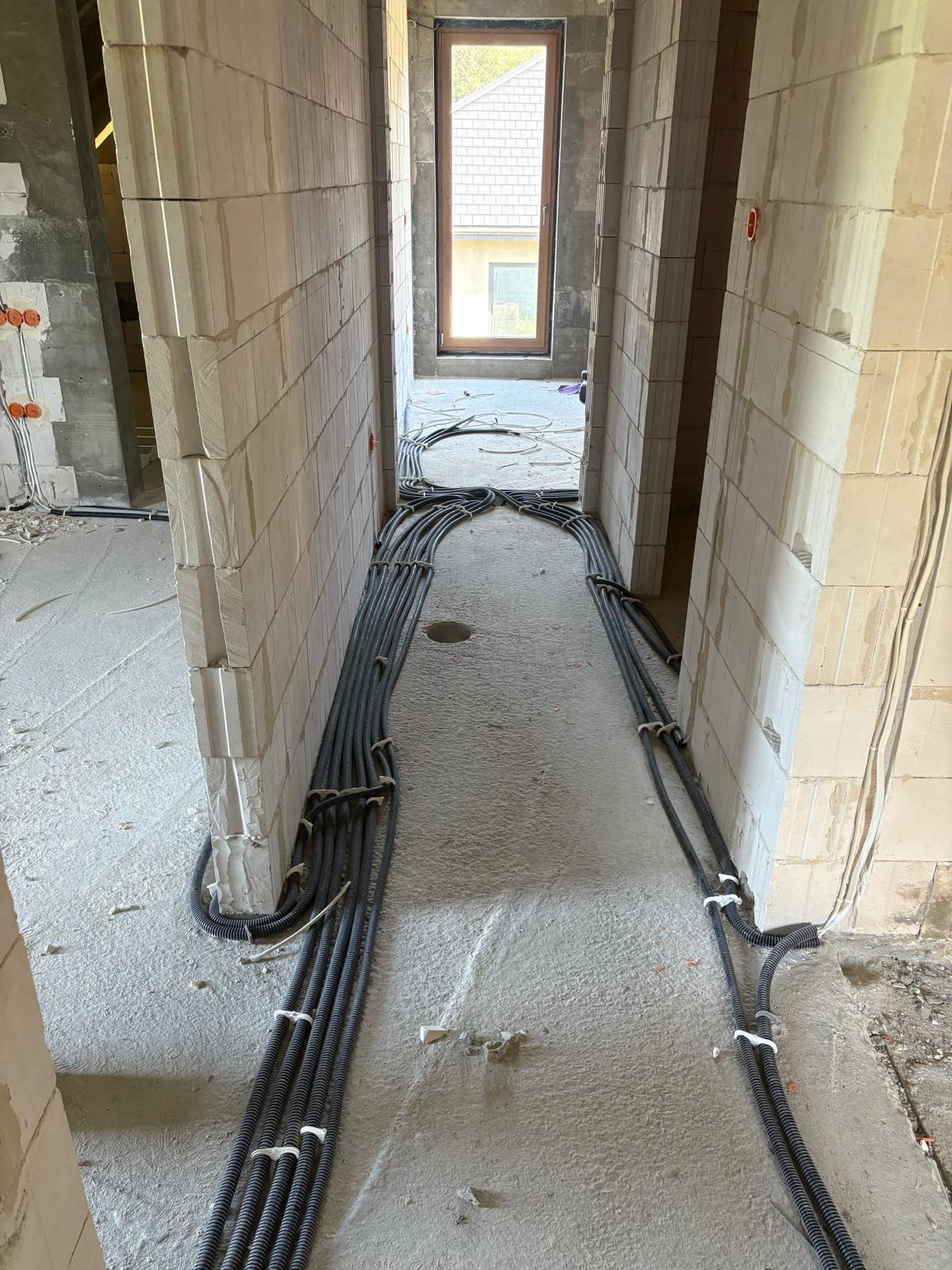 Korytarz w surowym stanie z ułożonymi przewodami w peszlach, przymocowanymi do podłoża białymi opaskami. Widoczne ściany z betonu komórkowego i okno w tle.