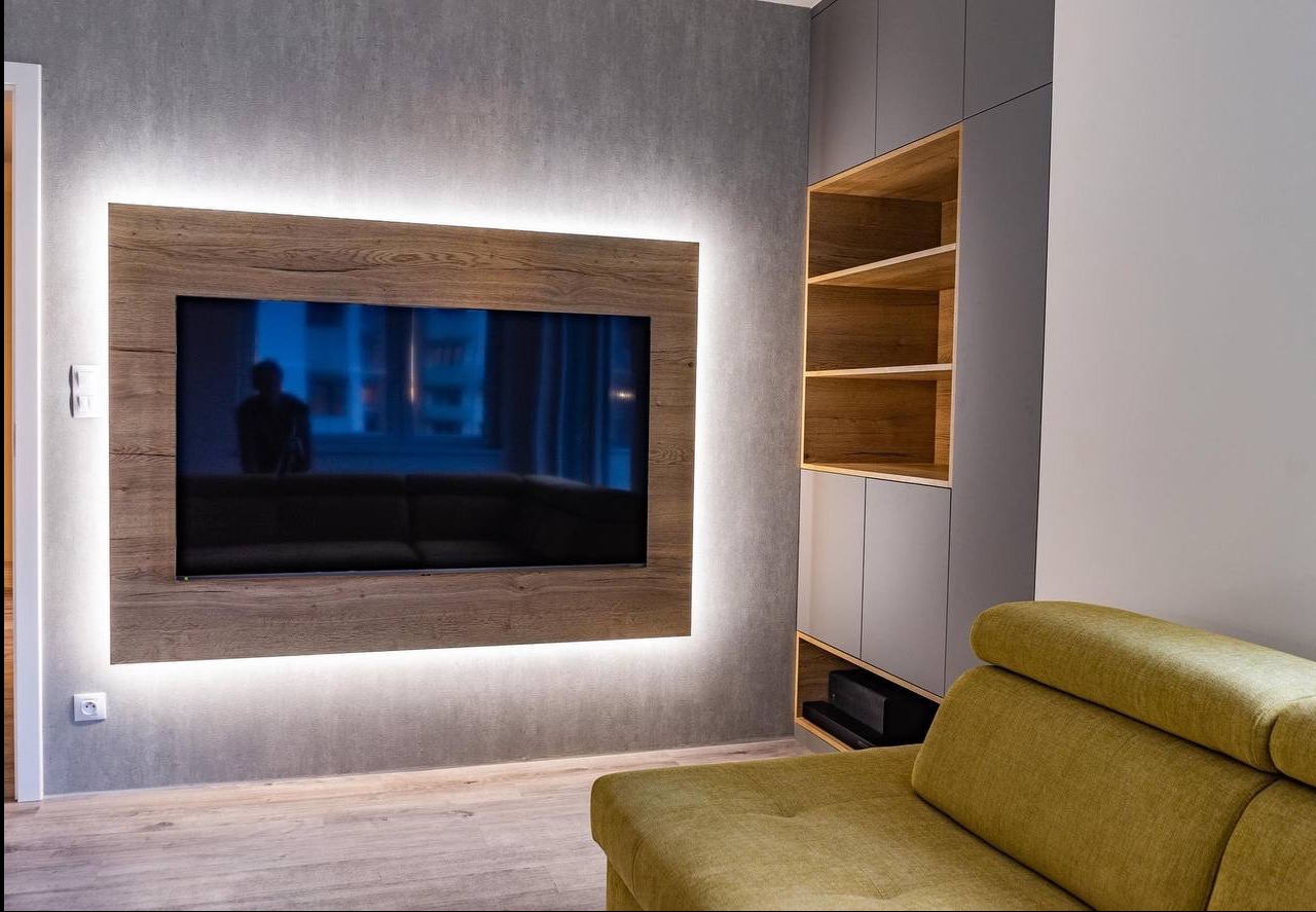 Nowoczesny salon z podświetlanym panelem TV w drewnianej ramie i szarą zabudową meblową z drewnianymi półkami. Częściowo widoczny narożnik w odcieniach zieleni.