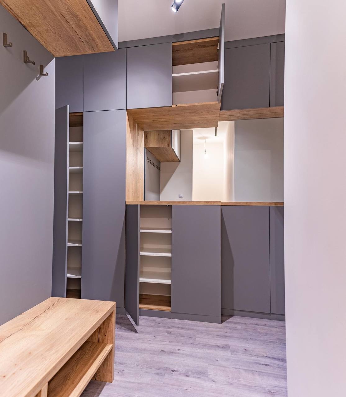 Szara zabudowa meblowa z drewnianymi akcentami w przedpokoju. Otwarta szafka z półkami, drewniana ławka. Nowoczesny design, minimalistyczny styl.