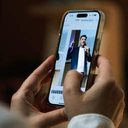 Jakub Groborz - Osoba trzyma telefon, na ekranie widoczny mężczyzna przemawiający do mikrofonu, prawdopodobnie podczas wydarzenia. Zdjęcie zrobione z bliska, skupia się na ekranie telefonu.