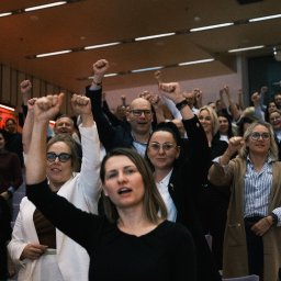 Jakub Groborz - Entuzjastyczny tłum z podniesionymi pięściami w sali konferencyjnej, oświetlonej ciepłym światłem. Uczestnicy w różnym wieku, z wyraźnymi emocjami na twarzach.