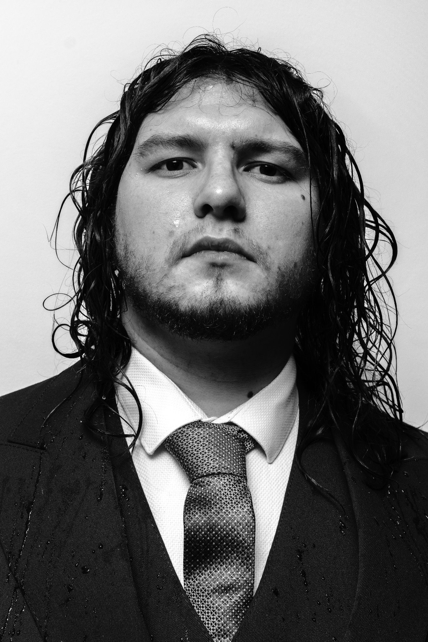 Czarno-biały portret mężczyzny w garniturze, z mokrymi włosami i kroplami wody na ubraniu, patrzącego prosto w obiektyw, w studio.