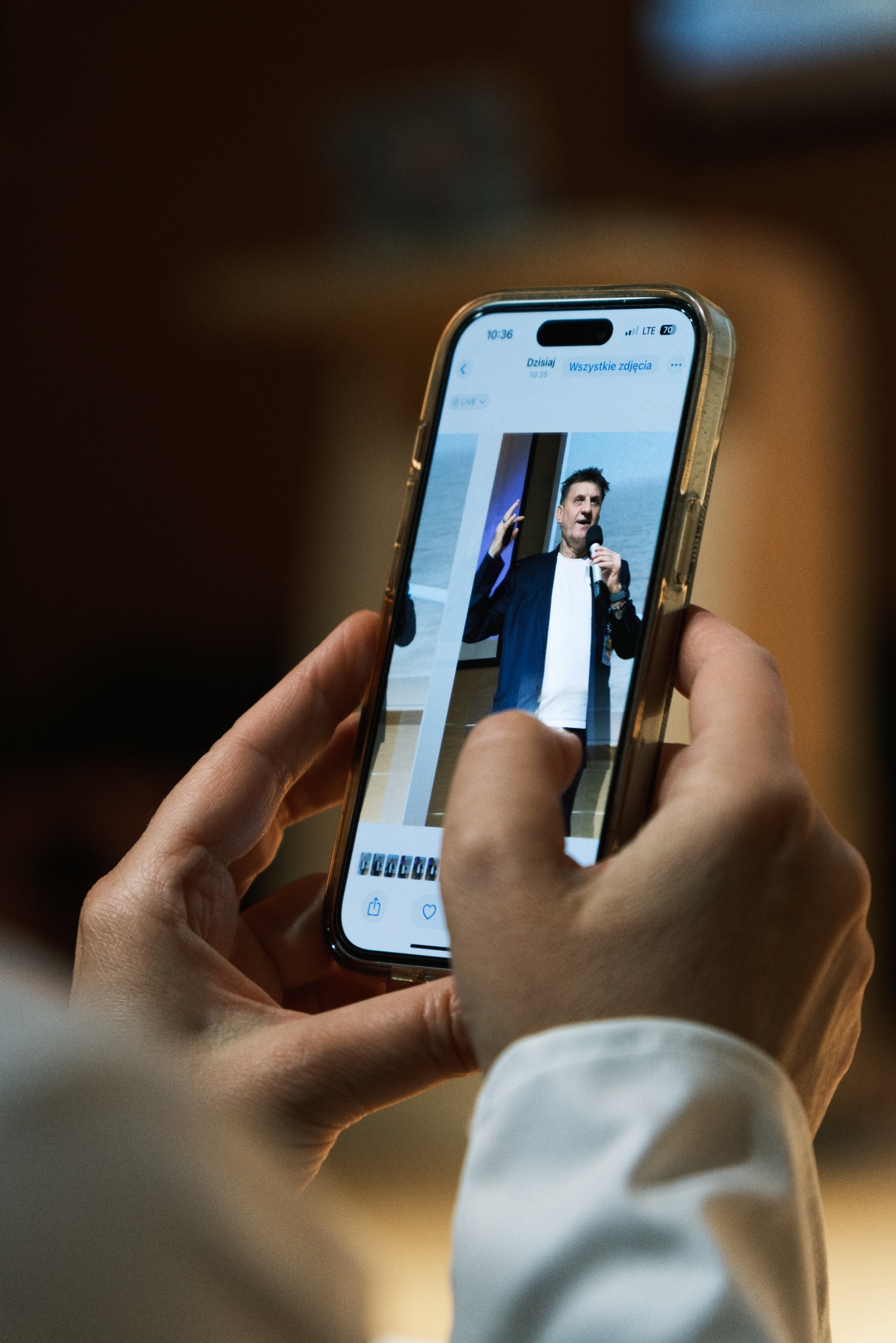 Osoba trzyma telefon, na ekranie widoczny mężczyzna przemawiający do mikrofonu, prawdopodobnie podczas wydarzenia. Zdjęcie zrobione z bliska, skupia się na ekranie telefonu.