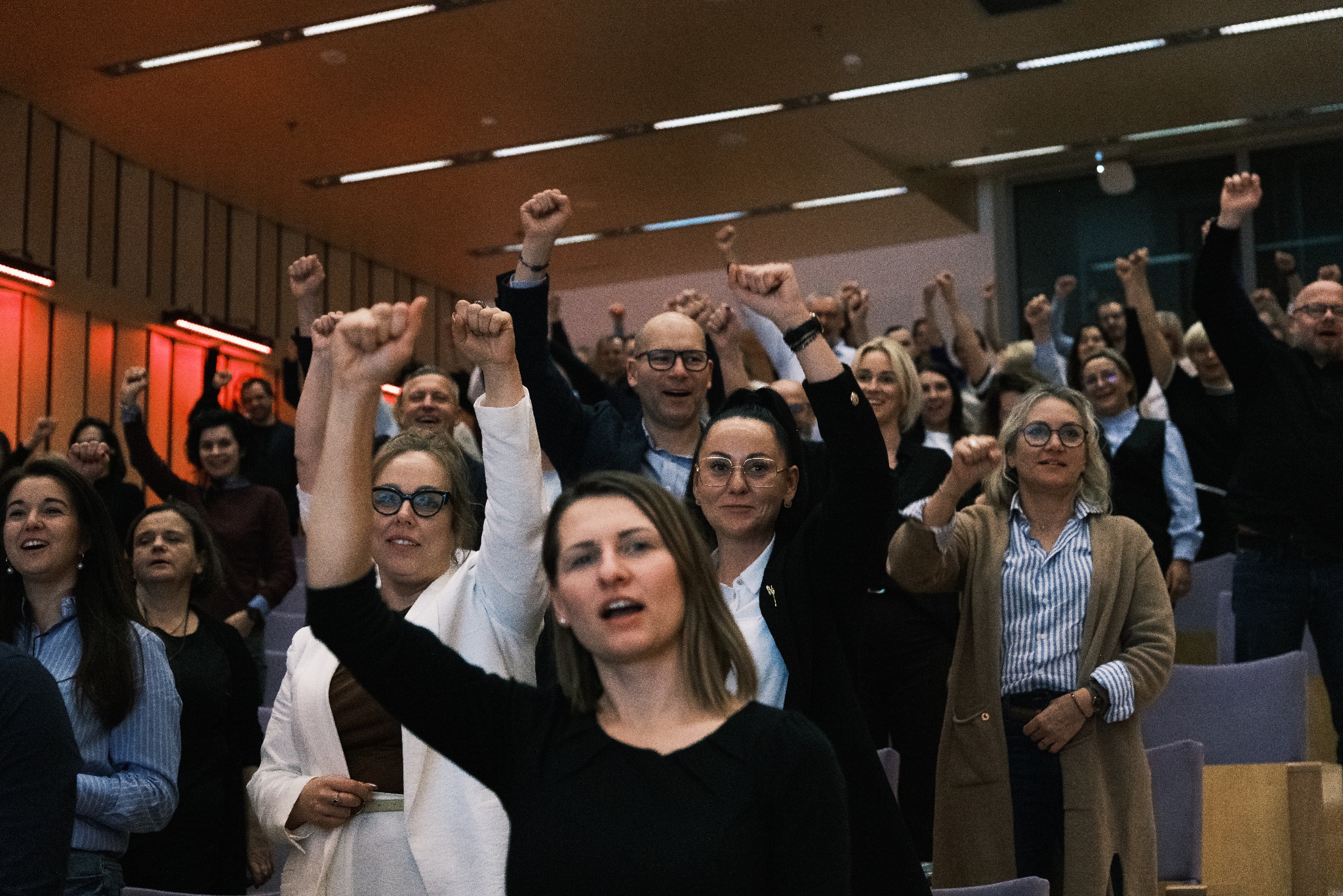 Entuzjastyczny tłum z podniesionymi pięściami w sali konferencyjnej, oświetlonej ciepłym światłem. Uczestnicy w różnym wieku, z wyraźnymi emocjami na twarzach.
