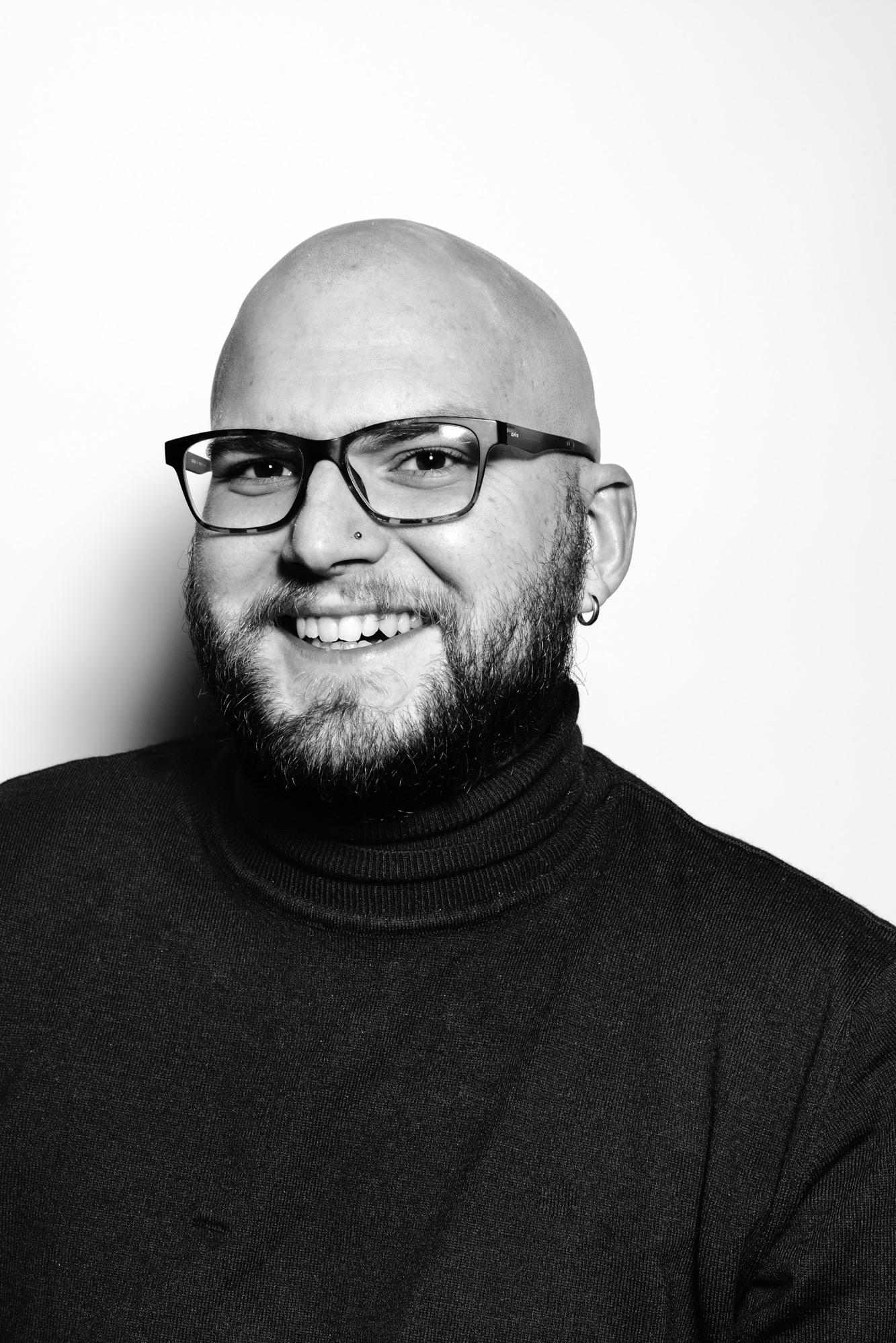 Czarno-biały portret uśmiechniętego mężczyzny w okularach i czarnym golfie na jasnym tle. Widoczne detale: kolczyk w uchu i piercing w nosie.