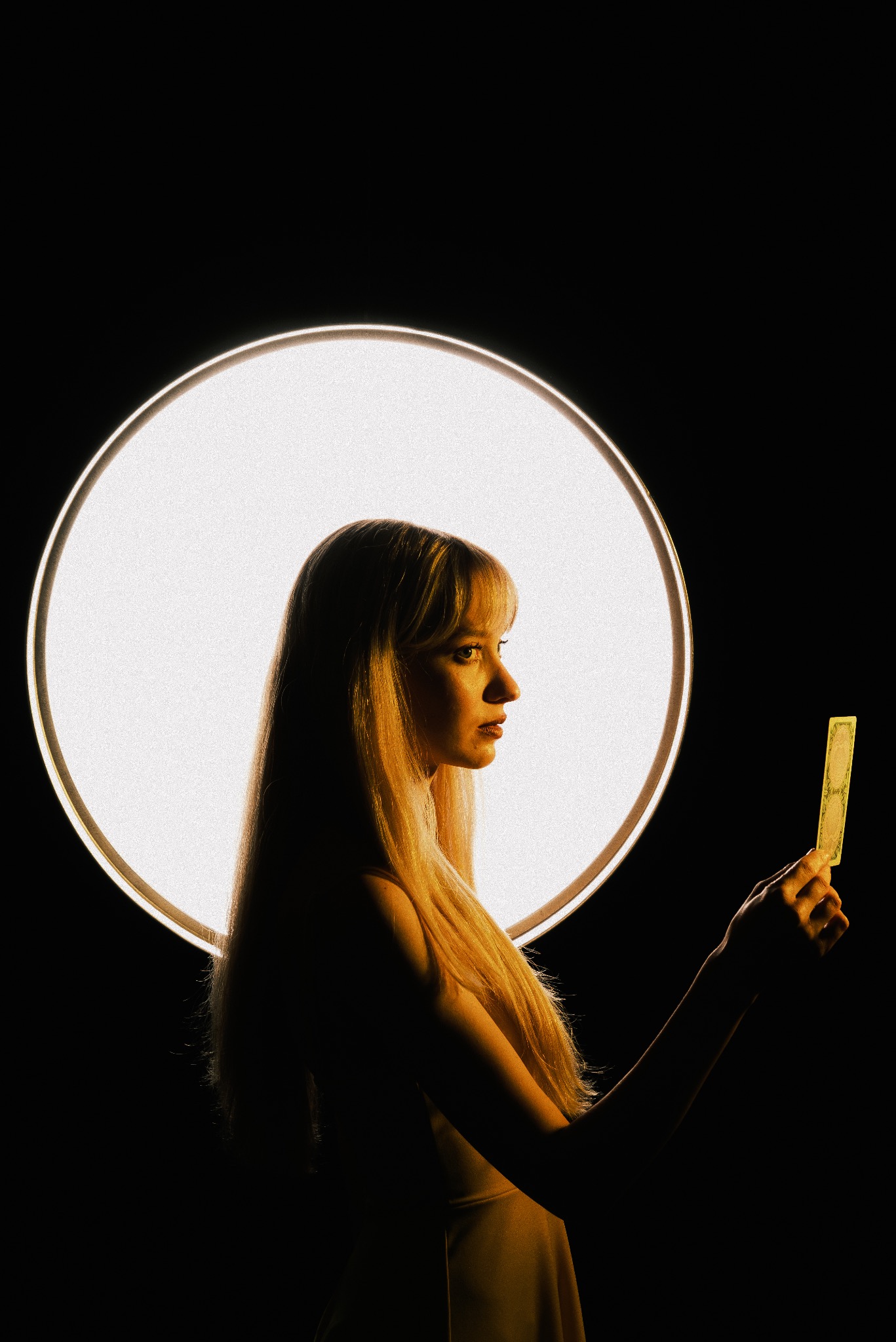 Profil kobiety z blond włosami, trzymającej kartę na tle okrągłego, jasnego światła. Artystyczne oświetlenie i ciemne tło tworzą unikalny portret.