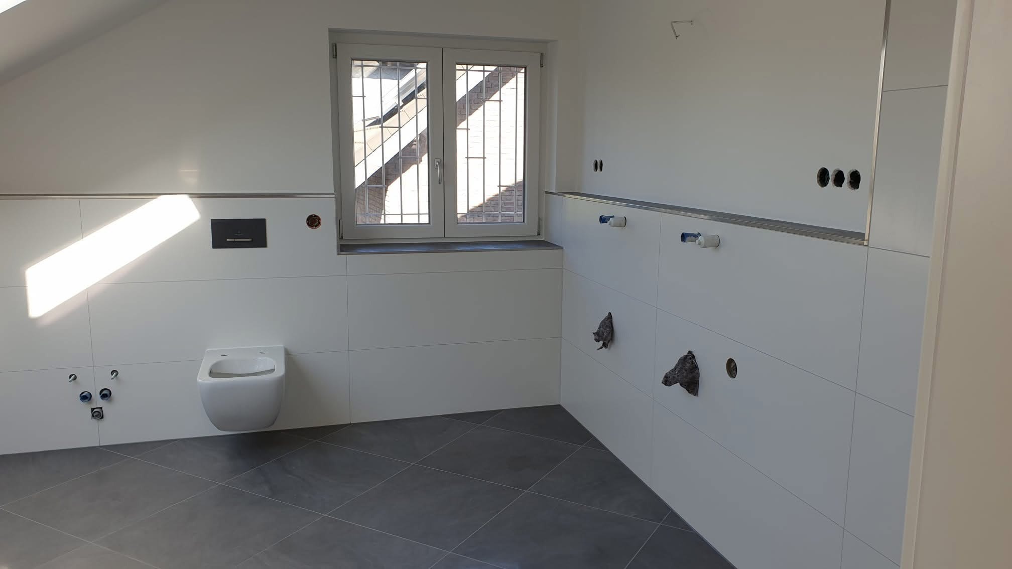 Biała, wykafelkowana łazienka w trakcie remontu. Widoczne przygotowane instalacje, podwieszana toaleta i okno z kratą. Szare płytki podłogowe.