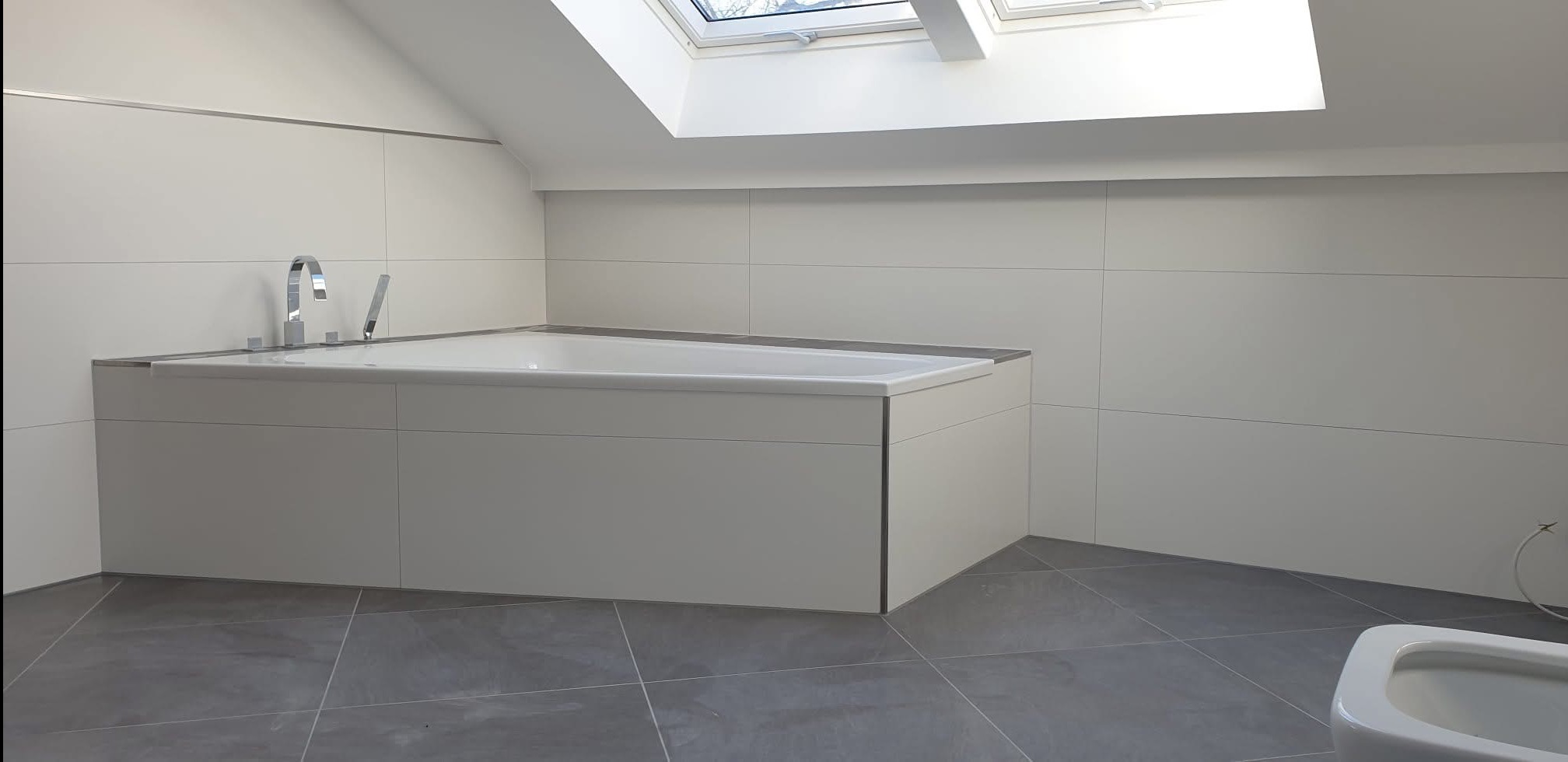 Nowoczesna łazienka na poddaszu z białą wanną obudowaną płytkami, szarą podłogą i oknem dachowym. Minimalistyczny design, chromowana bateria.