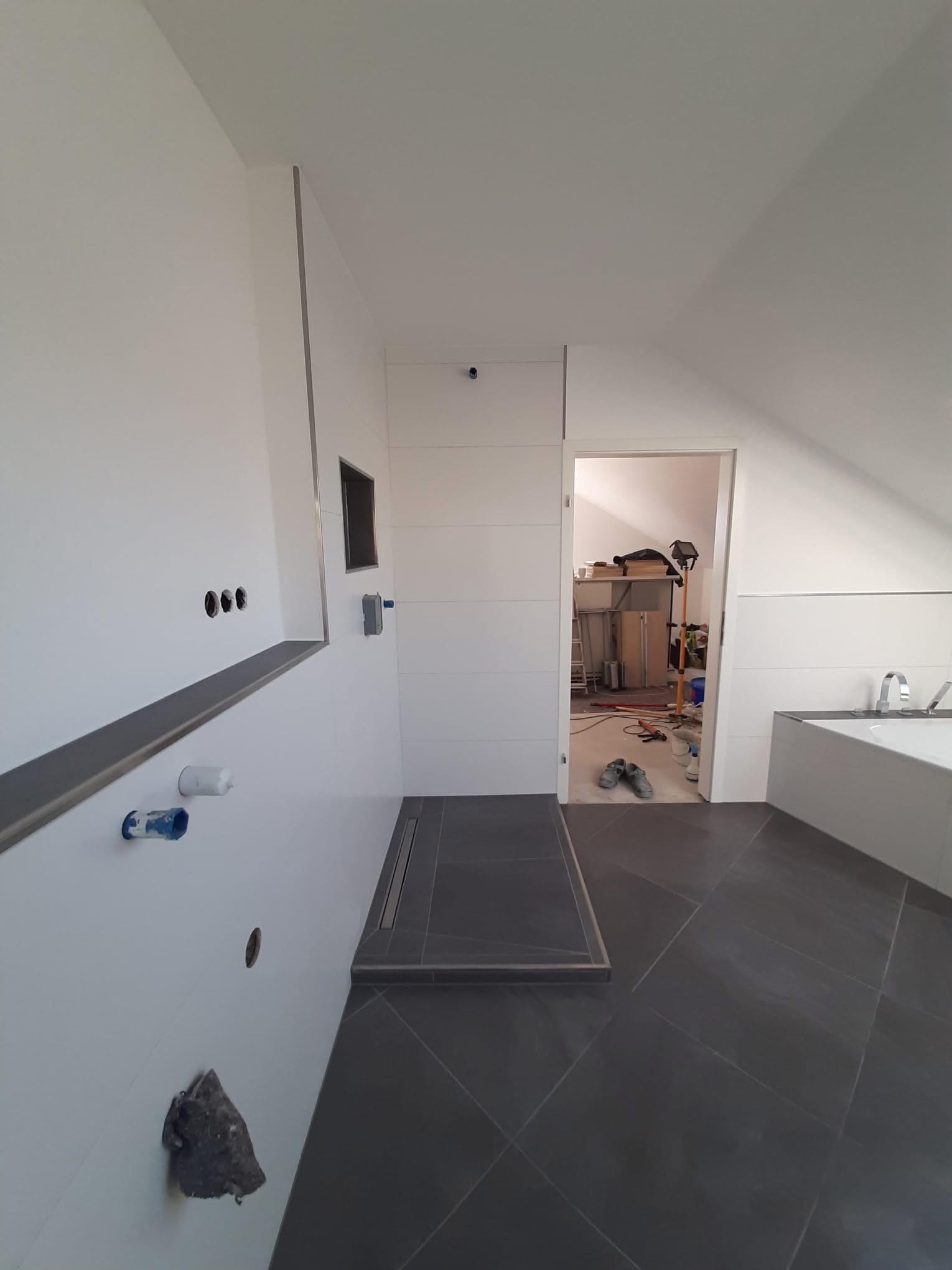 Łazienka w trakcie remontu: szare płytki podłogowe, białe ściany z przygotowanymi otworami na armaturę, widoczny odpływ liniowy i fragment wanny. W tle otwarte drzwi.