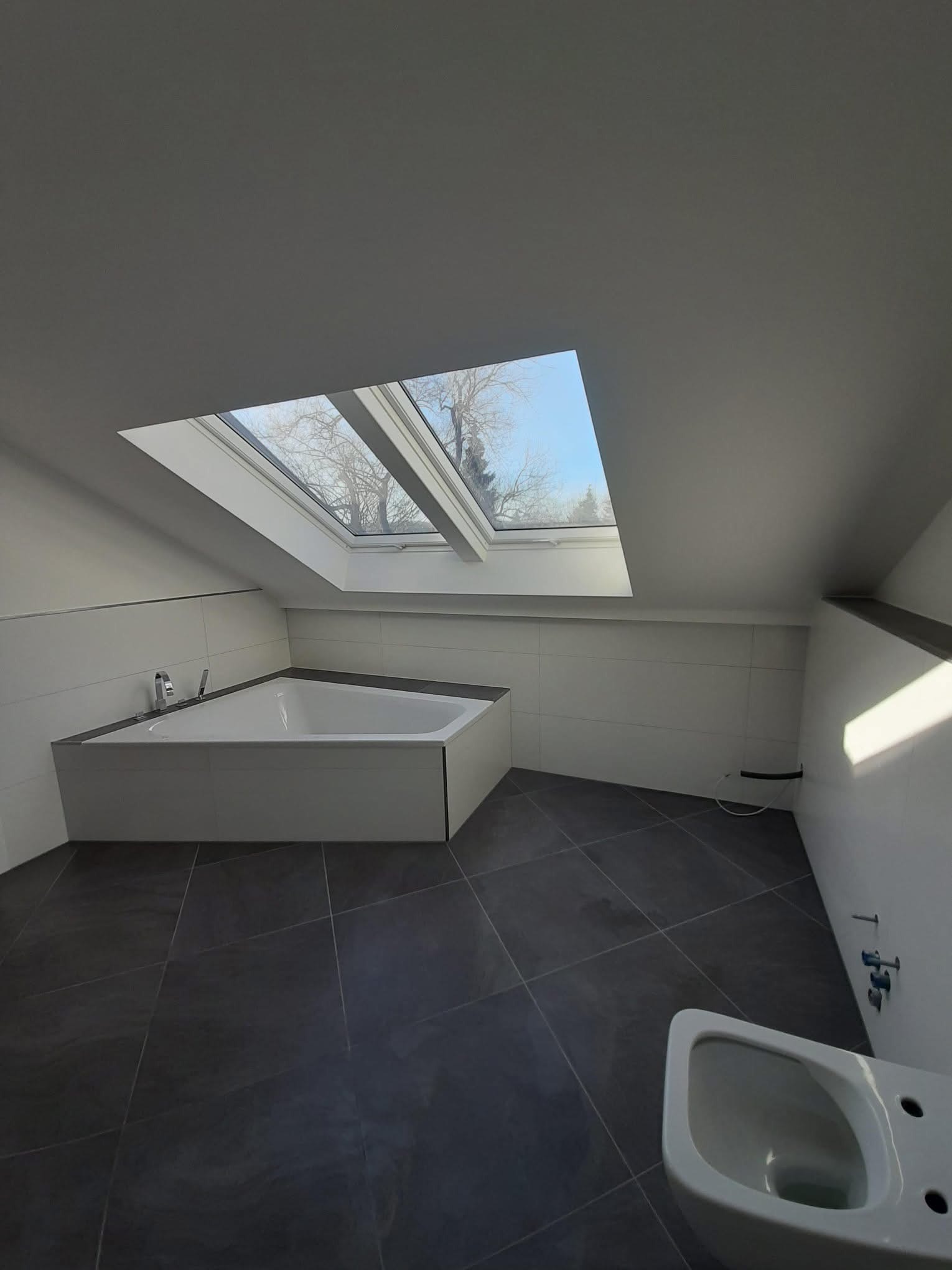 Nowoczesna łazienka na poddaszu z oknem dachowym, białą wanną narożną i ciemną podłogą z płytek. Minimalistyczny design, widok na drzewa przez okno.