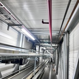 „Elektro-Jamboo” Dawid Jambor - Długi korytarz techniczny z widocznymi instalacjami pod sufitem: rury, kable w korytkach, oświetlenie. W oddali sylwetka osoby w białym kombinezonie.