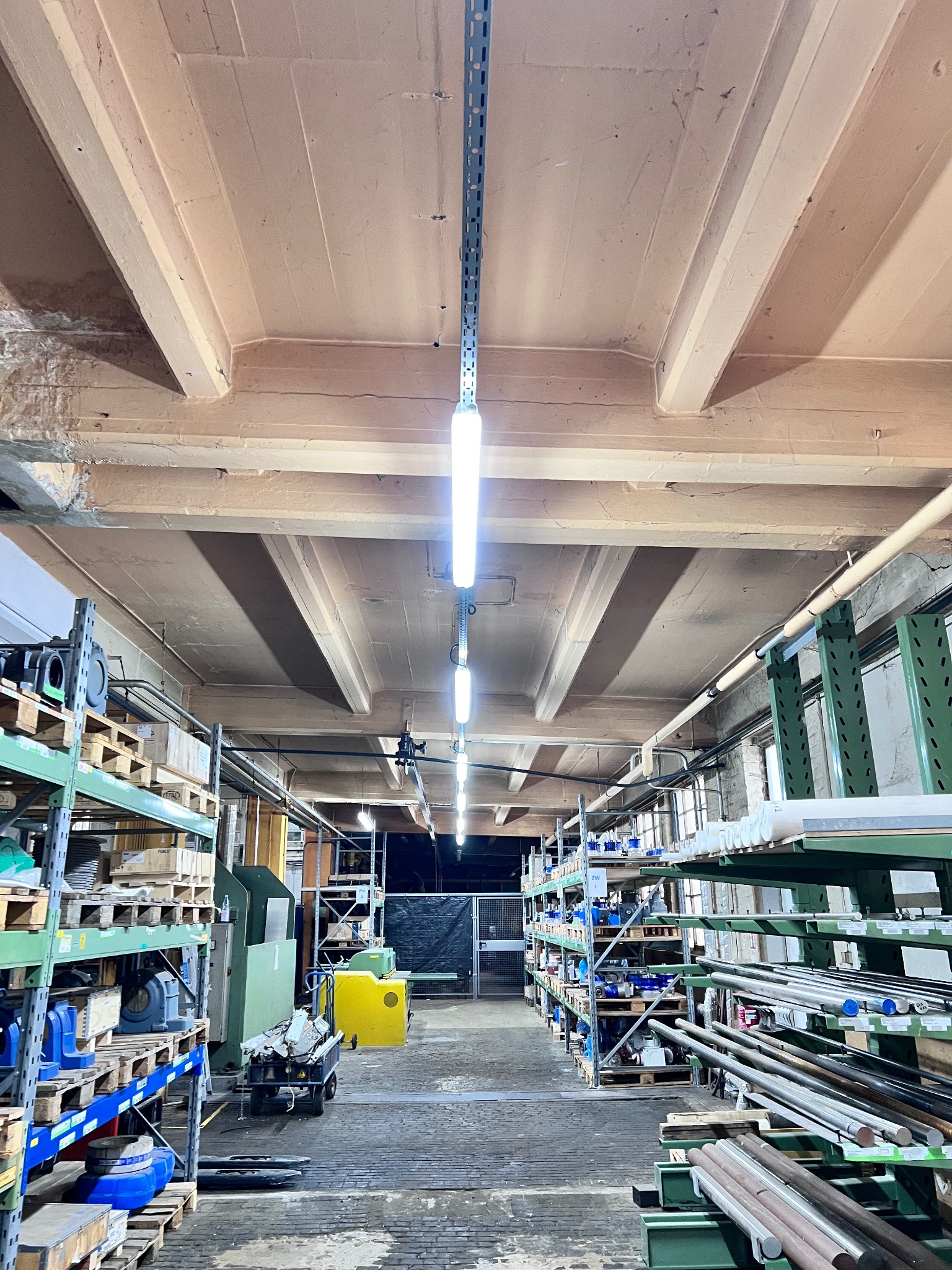 Długi magazyn z rzędami regałów wypełnionych materiałami. Na suficie widoczne lampy liniowe, oświetlające przestrzeń. Perspektywa z dołu, ukazująca konstrukcję sufitu.