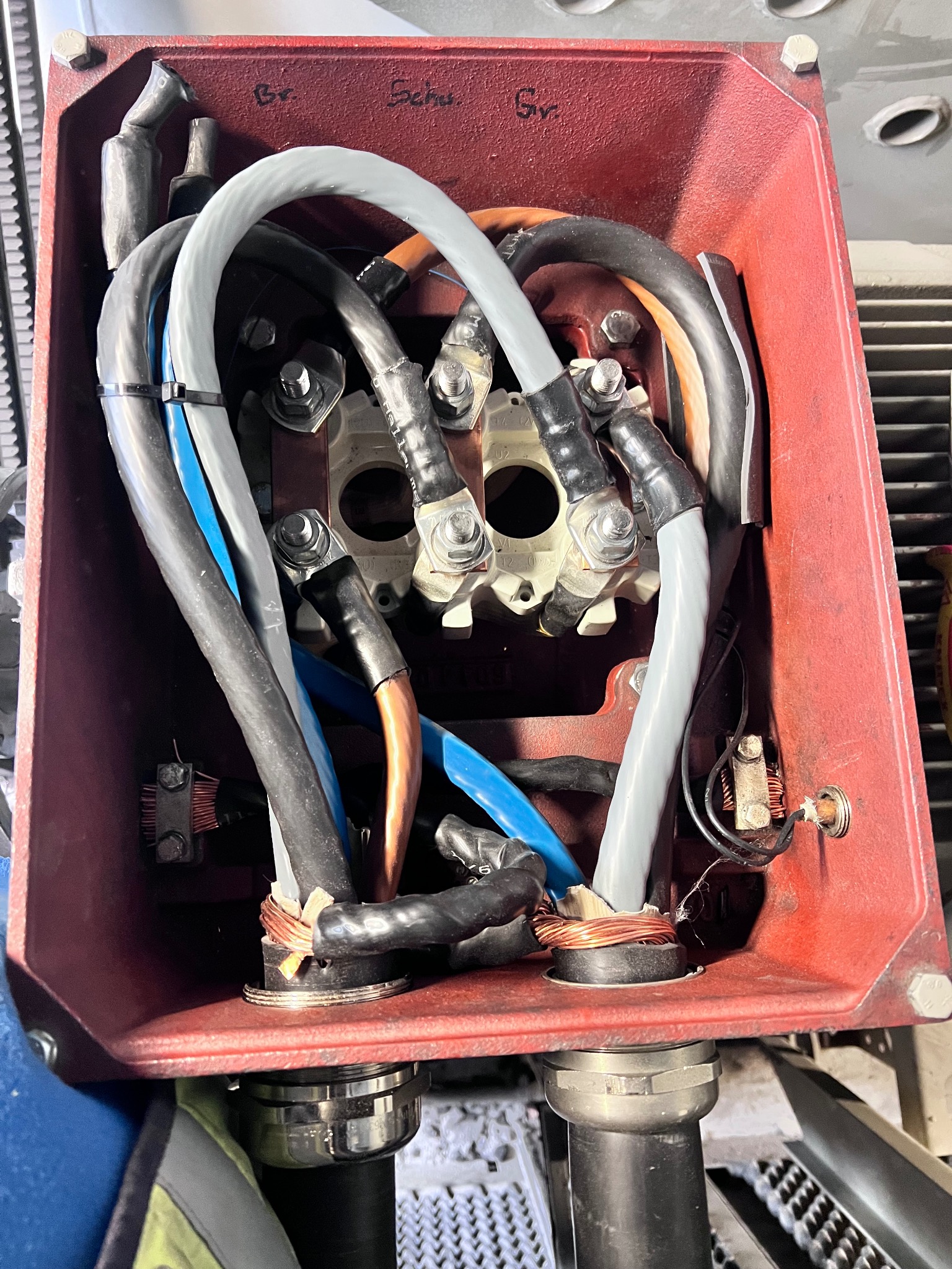 Wnętrze czerwonej skrzynki elektrycznej z grubymi kablami w izolacji szarej, czarnej i niebieskiej, podłączonymi do metalowych złączek. Widoczne ślady użytkowania i napisy na obudowie.