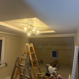 Wiktor Dominik Jagoda - Jasne wnętrze w trakcie prac wykończeniowych: drabina malarska, lampa sufitowa, świeżo pomalowany sufit z dekoracyjnym wgłębieniem i oświetleniem. Widoczne narzędzia i materiały.