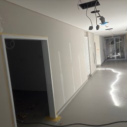 Wiktor Dominik Jagoda - Korytarz w trakcie prac wykończeniowych: szpachlowane ściany, zabezpieczone taśmą framugi drzwi, widoczne przewody oświetleniowe. Podłoga pokryta świeżą wylewką.