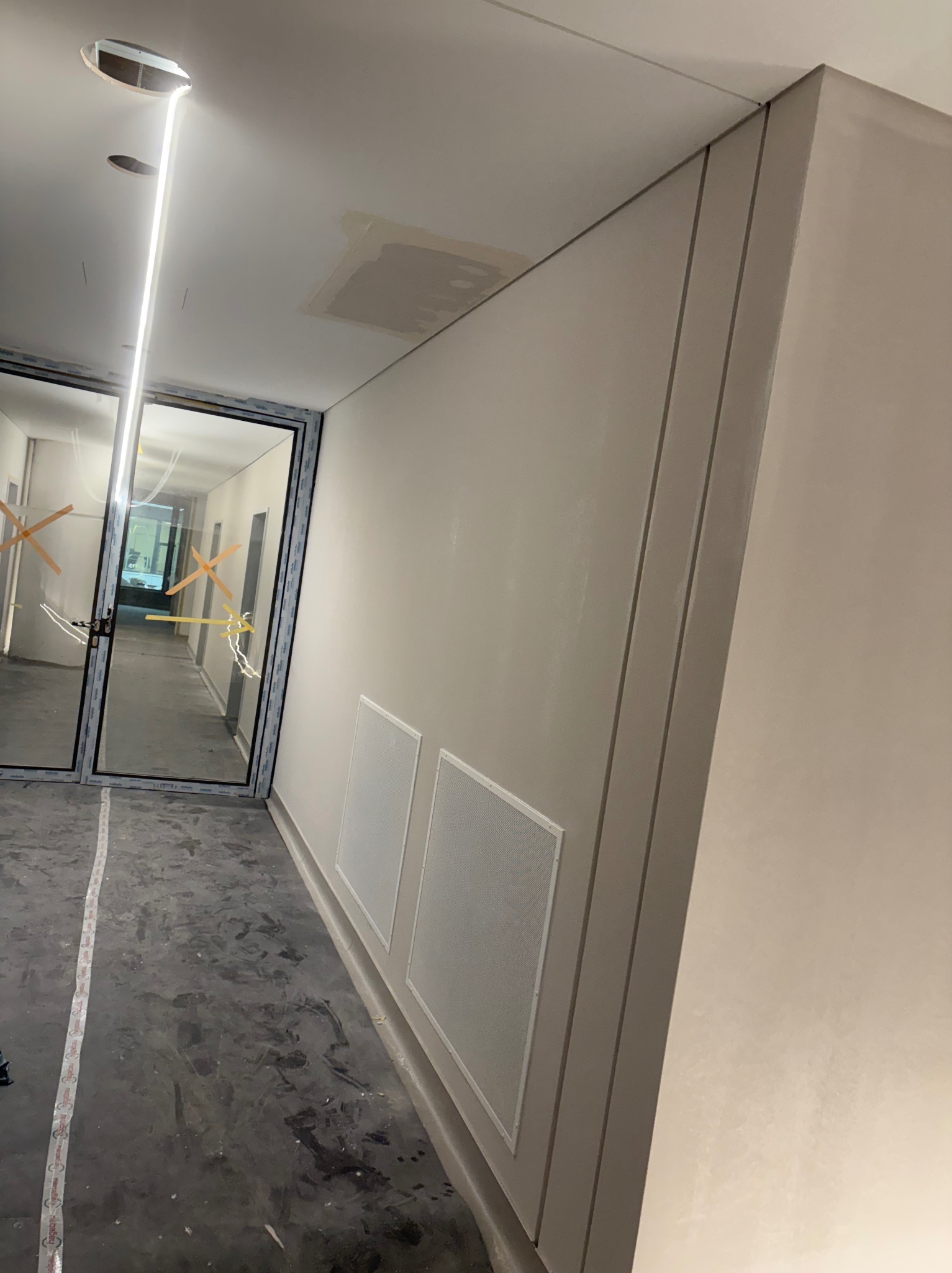 Korytarz w trakcie prac wykończeniowych: ściany po szpachlowaniu, widoczne otwory w suficie i zabezpieczone taśmą drzwi. Podłoga zabezpieczona folią.