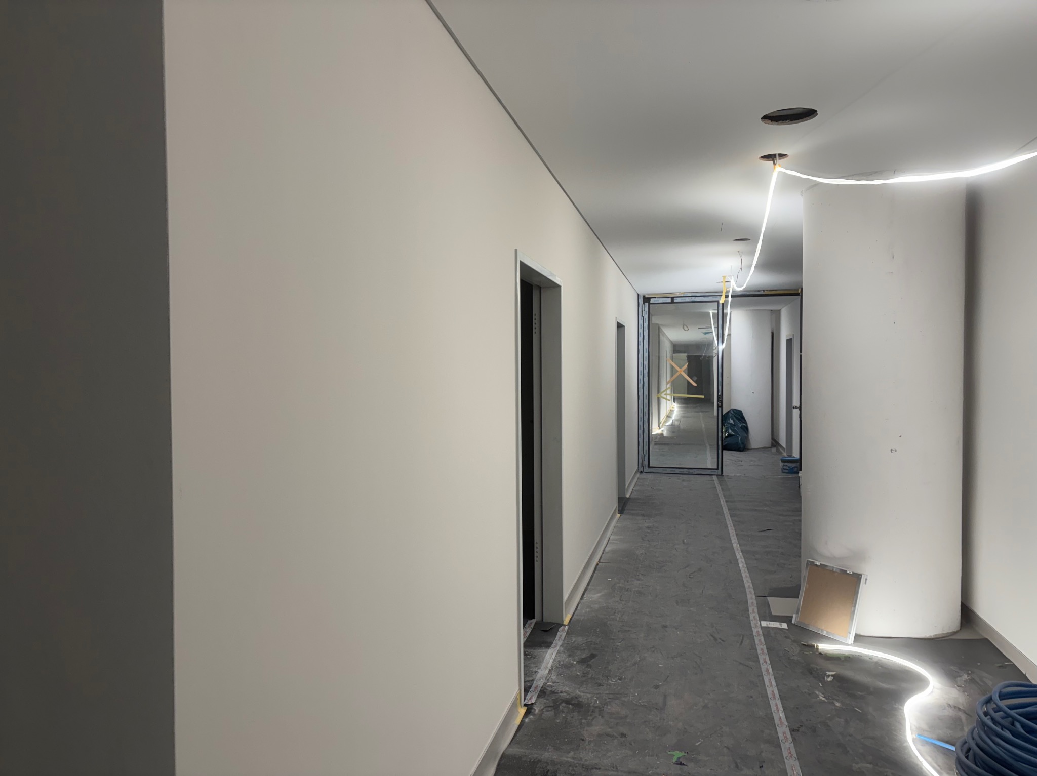 Korytarz w trakcie prac wykończeniowych: świeżo pomalowane ściany w odcieniach bieli, szara podłoga, widoczne instalacje elektryczne i materiały budowlane.