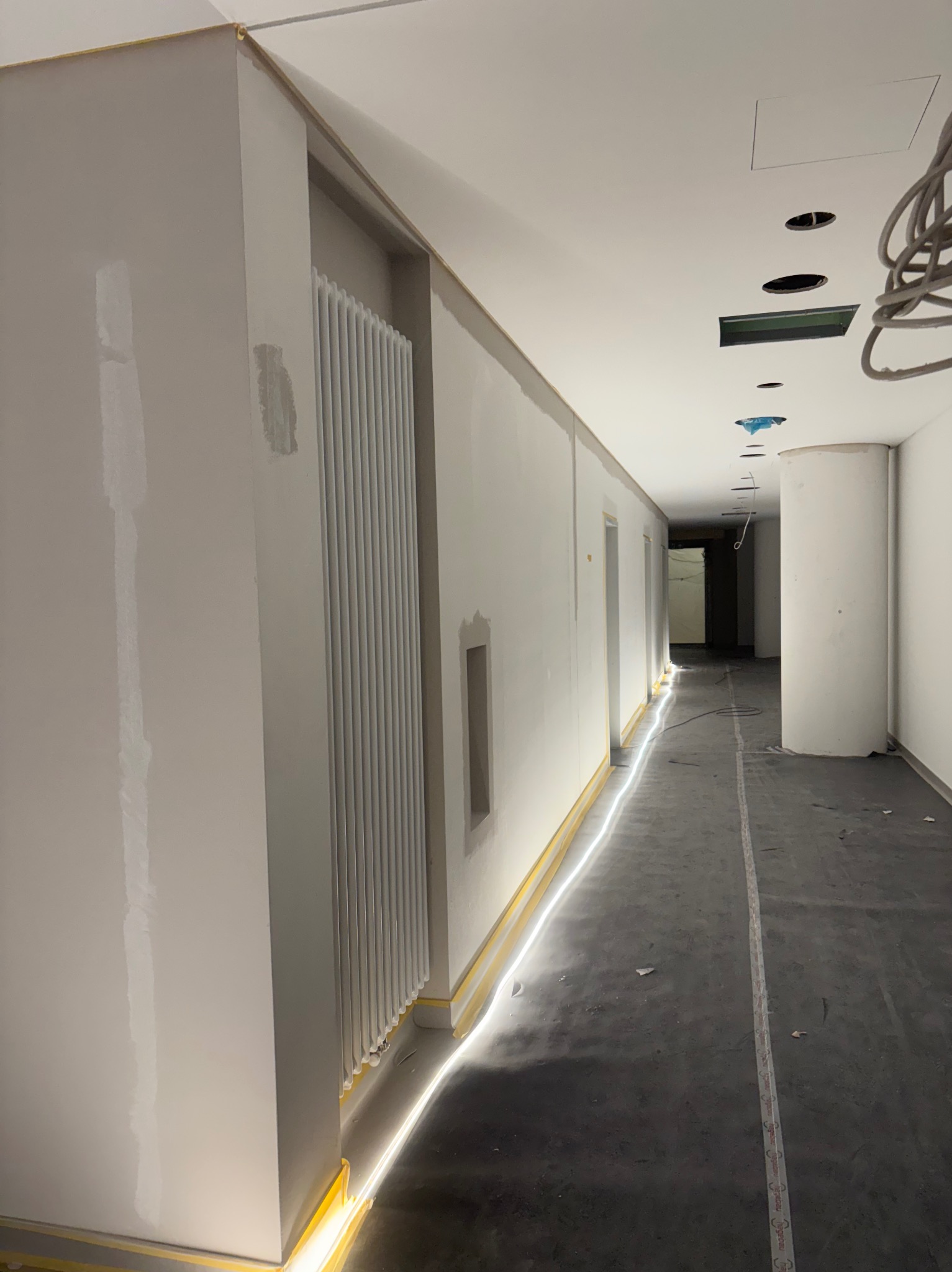 Korytarz w trakcie prac wykończeniowych: ściany po szpachlowaniu, zabezpieczone taśmą malarską, grzejnik, sufit z otworami na oświetlenie. Widok w głąb korytarza.