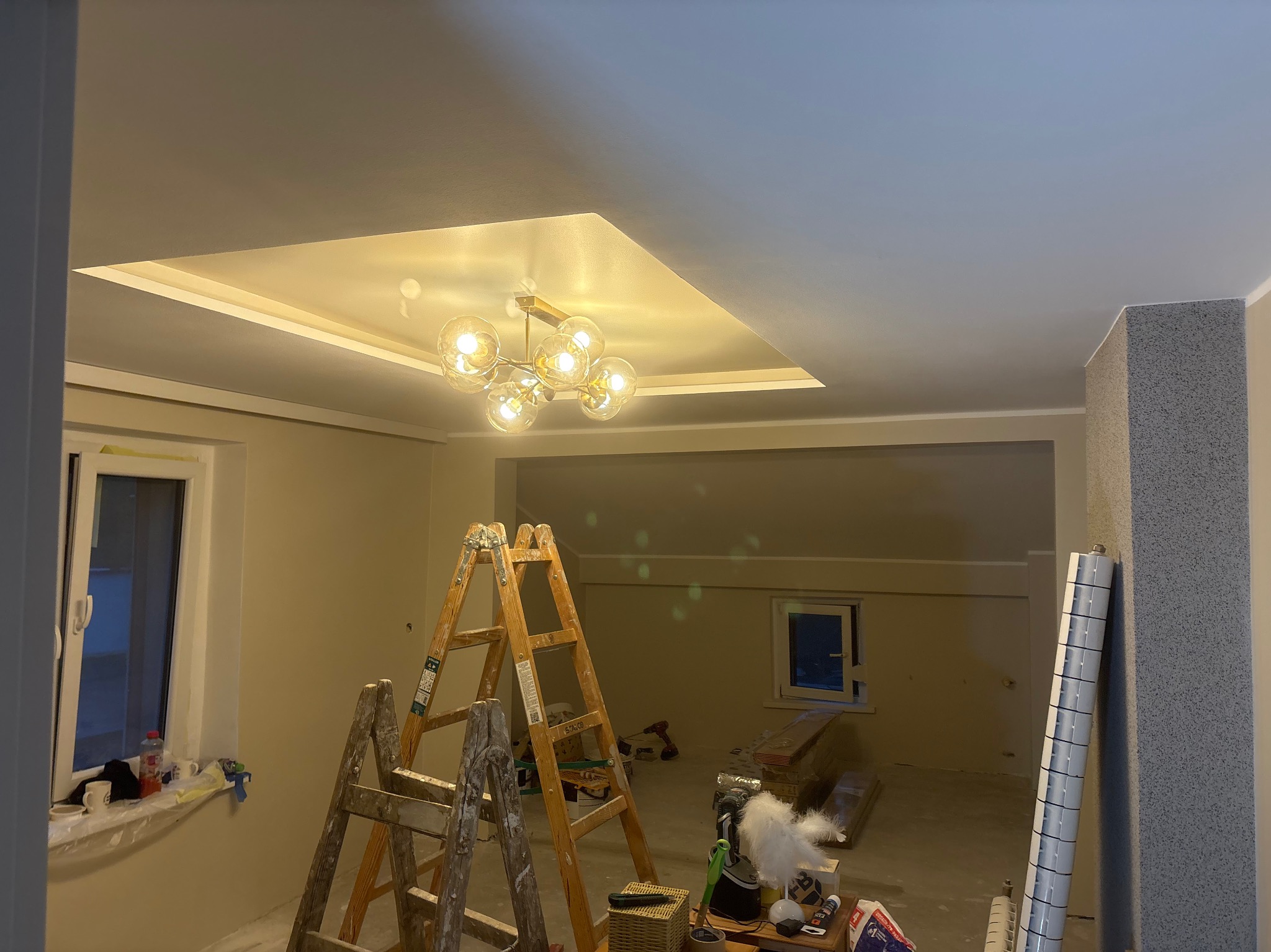 Jasne wnętrze w trakcie prac wykończeniowych: drabina malarska, lampa sufitowa, świeżo pomalowany sufit z dekoracyjnym wgłębieniem i oświetleniem. Widoczne narzędzia i materiały.