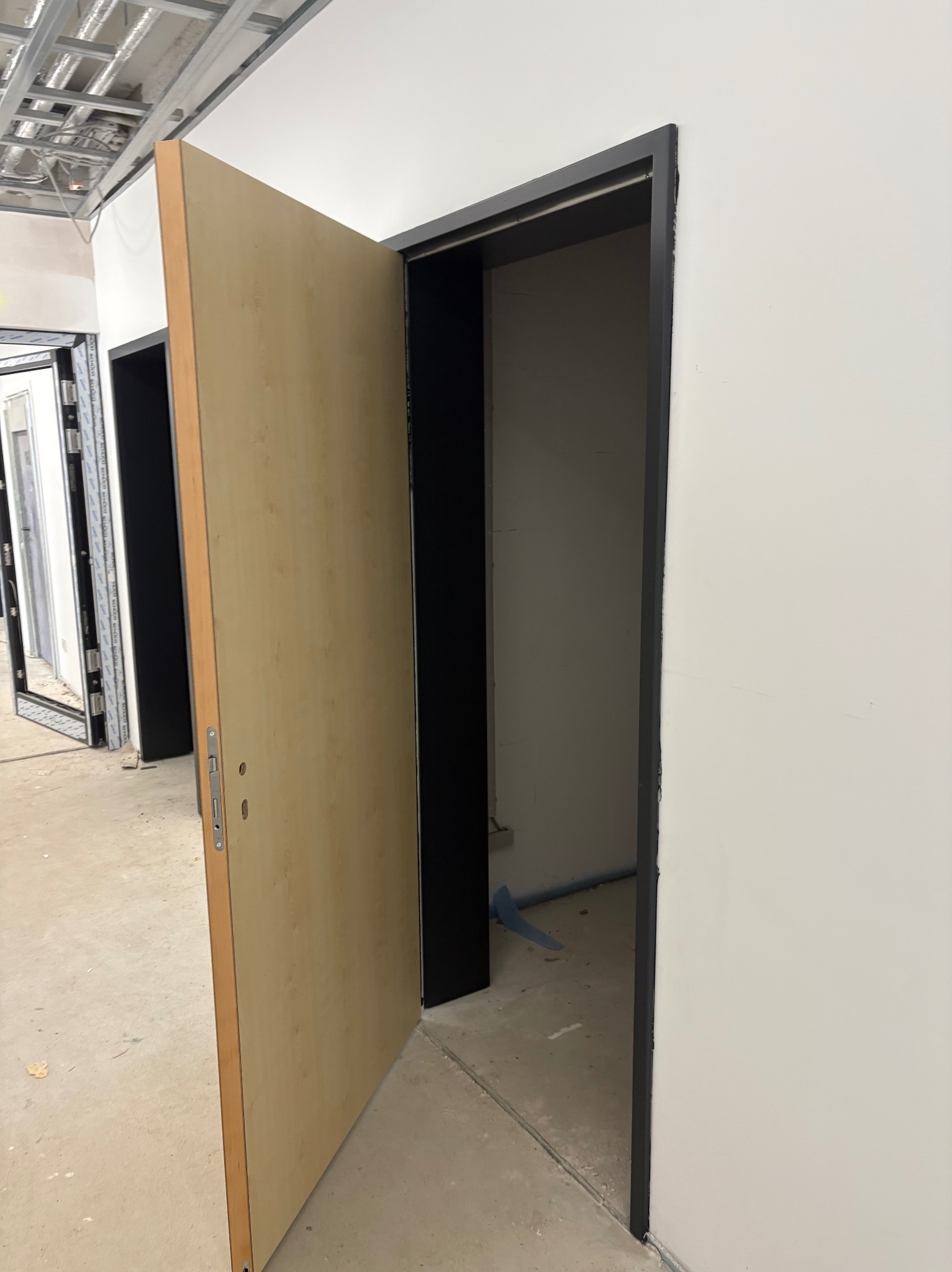 Otwarta, jasna, drewniana płycinowa drzwi z czarną ościeżnicą na tle świeżo pomalowanej białej ściany. Widoczny fragment sufitu podwieszanego i inne drzwi w tle.