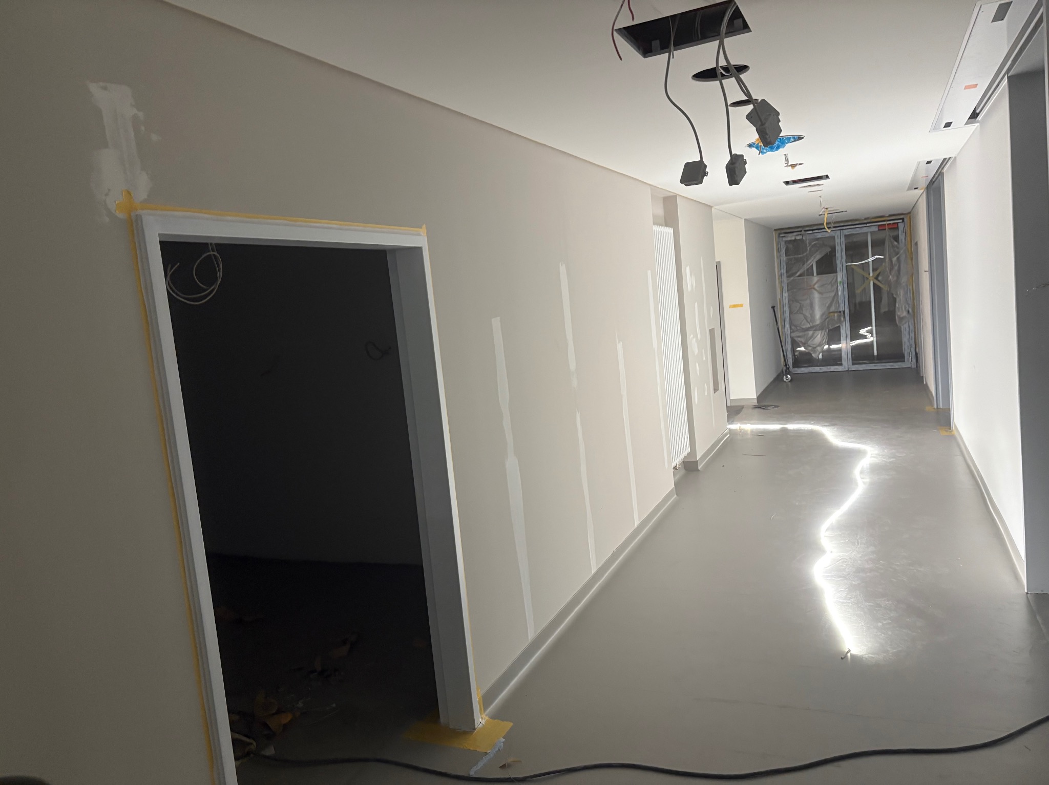 Korytarz w trakcie prac wykończeniowych: szpachlowane ściany, zabezpieczone taśmą framugi drzwi, widoczne przewody oświetleniowe. Podłoga pokryta świeżą wylewką.