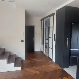 Oleh Mytulynskyi - Nowoczesne wykończenie wnętrza w Oławie: drewniana podłoga w jodełkę, schody z zabudową, czarna zabudowa kuchenna i przesuwne drzwi ze szprosami.