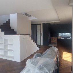 Oleh Mytulynskyi - Nowoczesne wnętrze z drewnianą podłogą w jodełkę, białymi schodami z wbudowanymi półkami i czarną kuchnią w tle. Sofa zabezpieczona folią ochronną.
