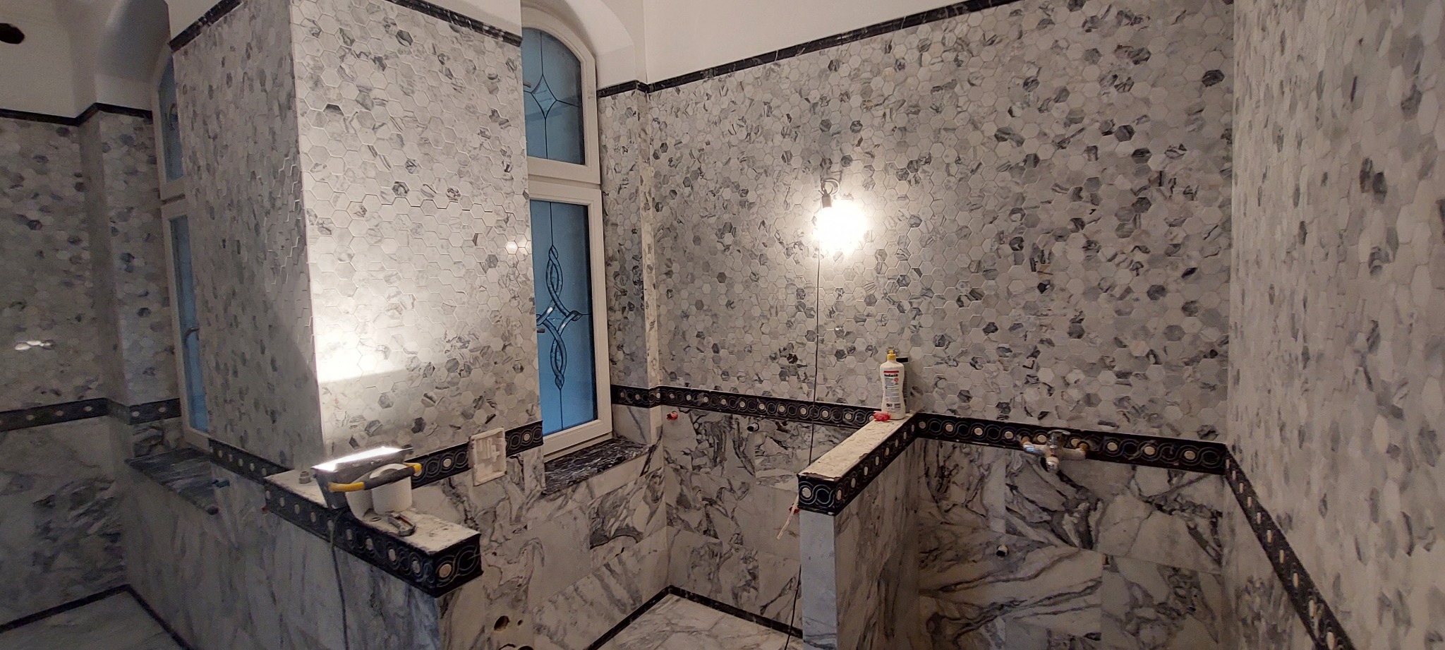 Mozaika heksagonalna w odcieniach szarości zdobi ściany łazienki z oknem. Dekoracyjne listwy i marmurowe wykończenie dodają elegancji. Widoczne narzędzia i chemia budowlana.