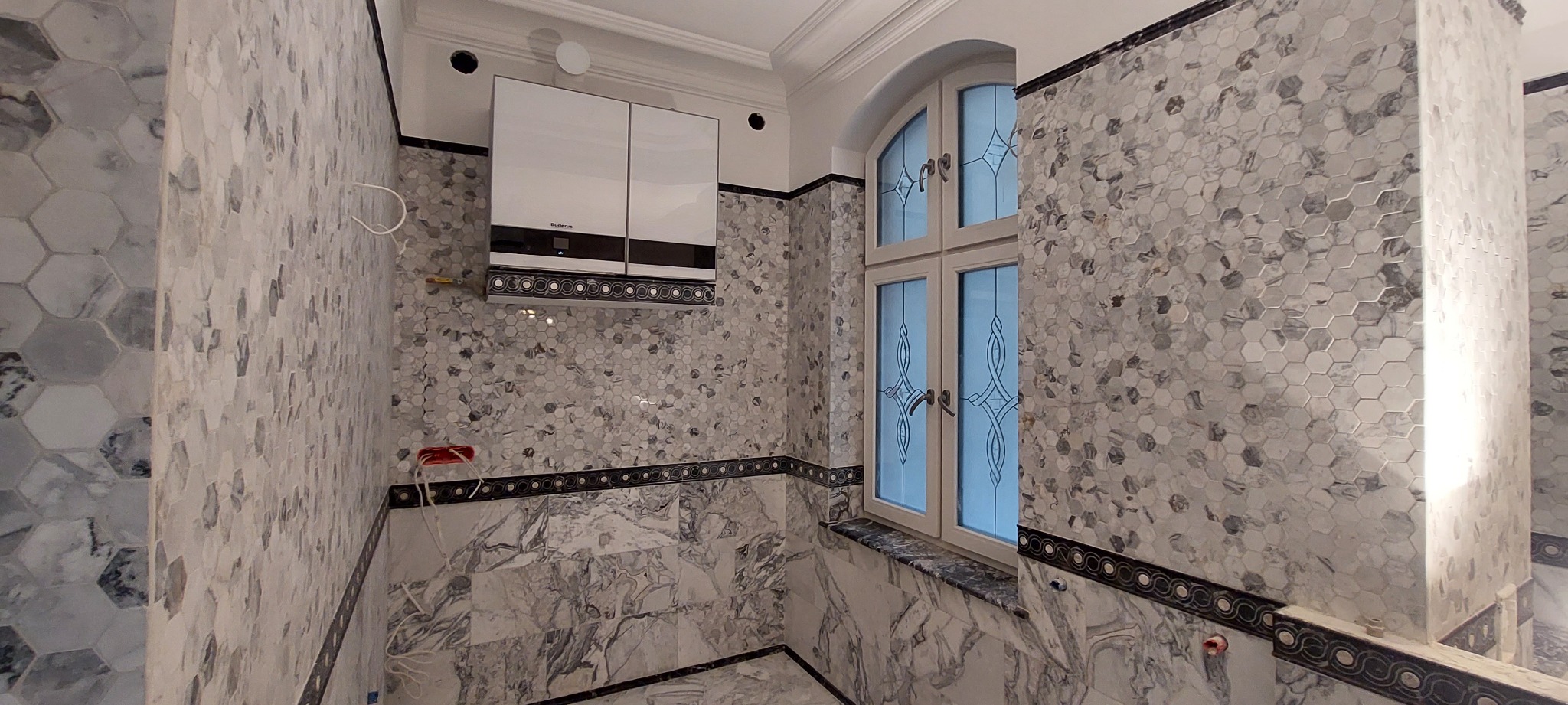 Wykończona łazienka z heksagonalnymi płytkami w odcieniach szarości i bieli, dekoracyjnym pasem płytek i oknem z ozdobnymi szprosami. Widoczny piec gazowy.