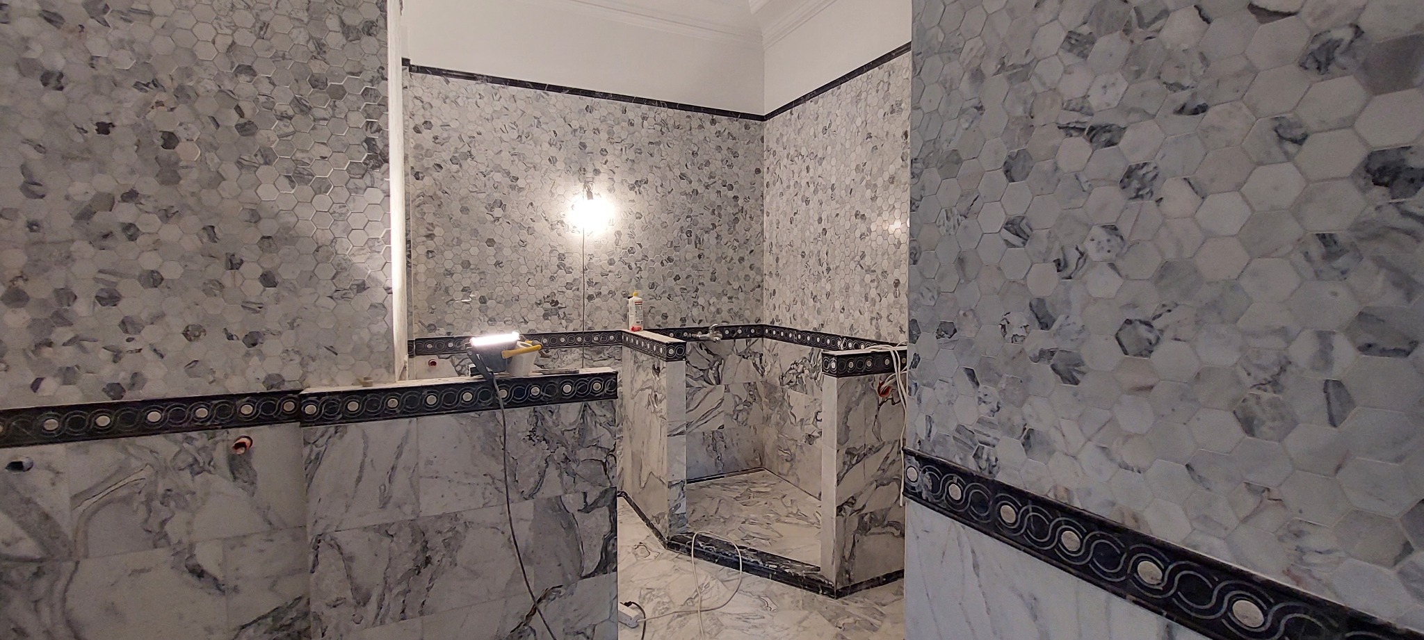 Łazienka w trakcie wykańczania: marmurowe płytki heksagonalne na ścianach i podłodze, dekoracyjny pas z motywem fal, oświetlenie robocze, widoczne przewody.