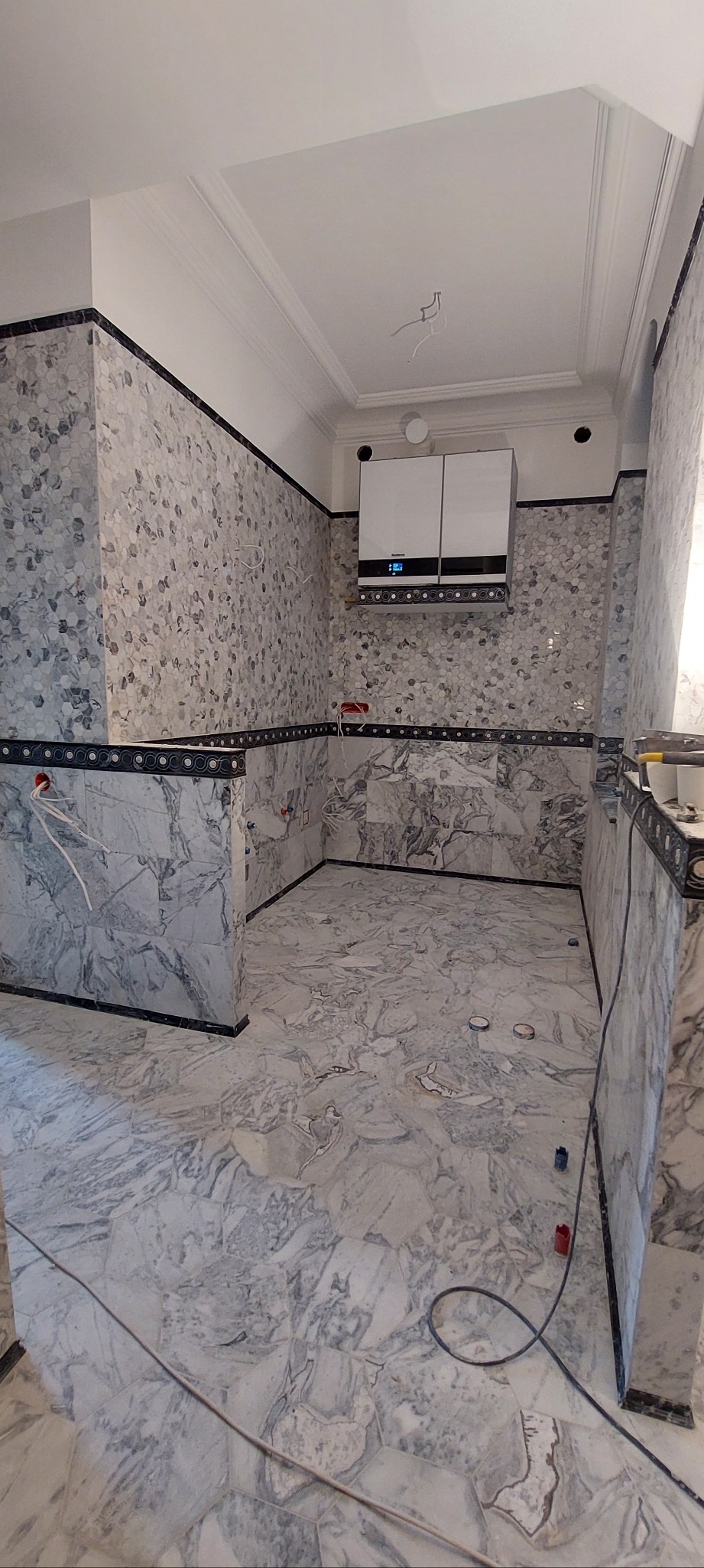 Wykończona łazienka z marmurowymi płytkami na ścianach i podłodze, z zamontowanym piecem gazowym. Widoczne narzędzia glazurnicze.