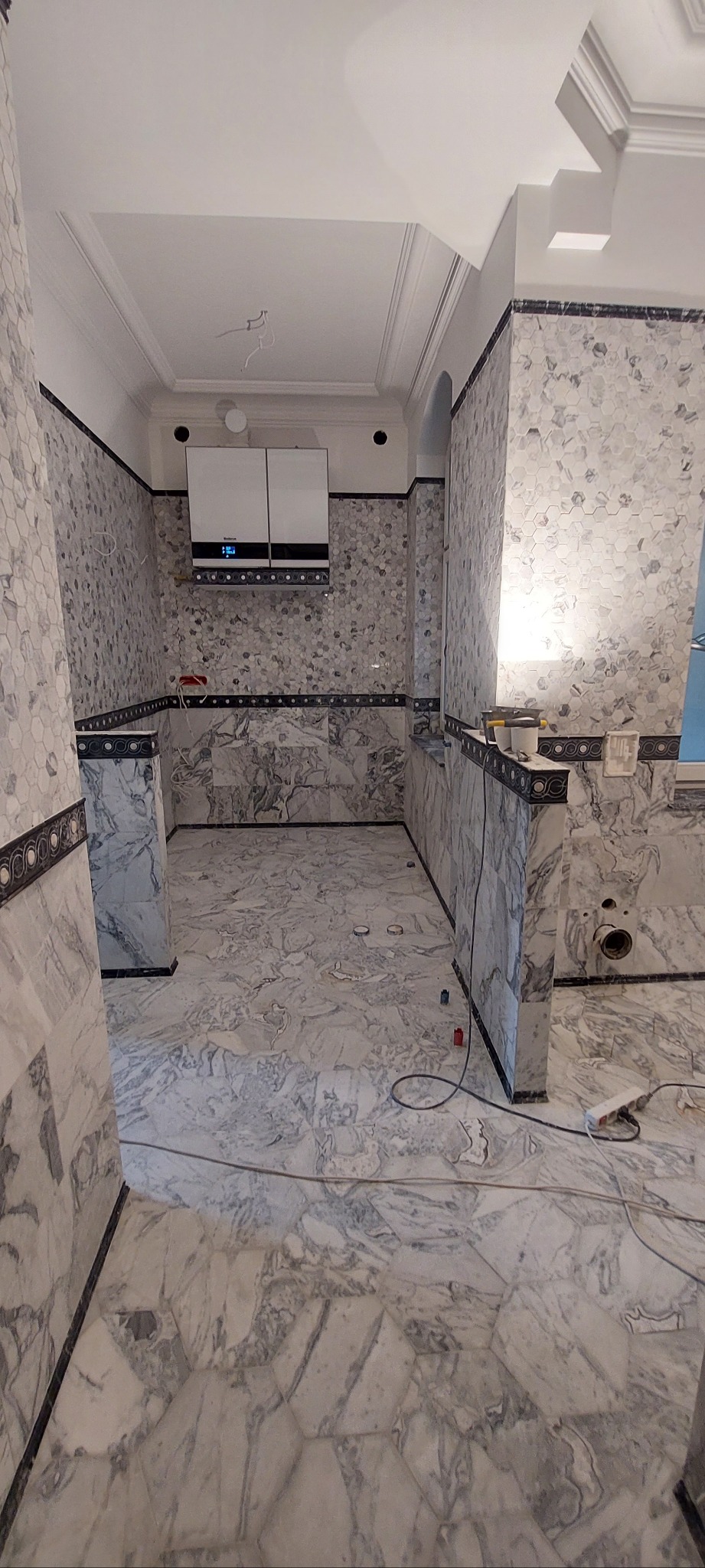 Wykończona łazienka z marmurowymi płytkami heksagonalnymi na ścianach i podłodze. Widoczny piec dwufunkcyjny. Dekoracyjne listwy. Częściowo widoczne narzędzia.